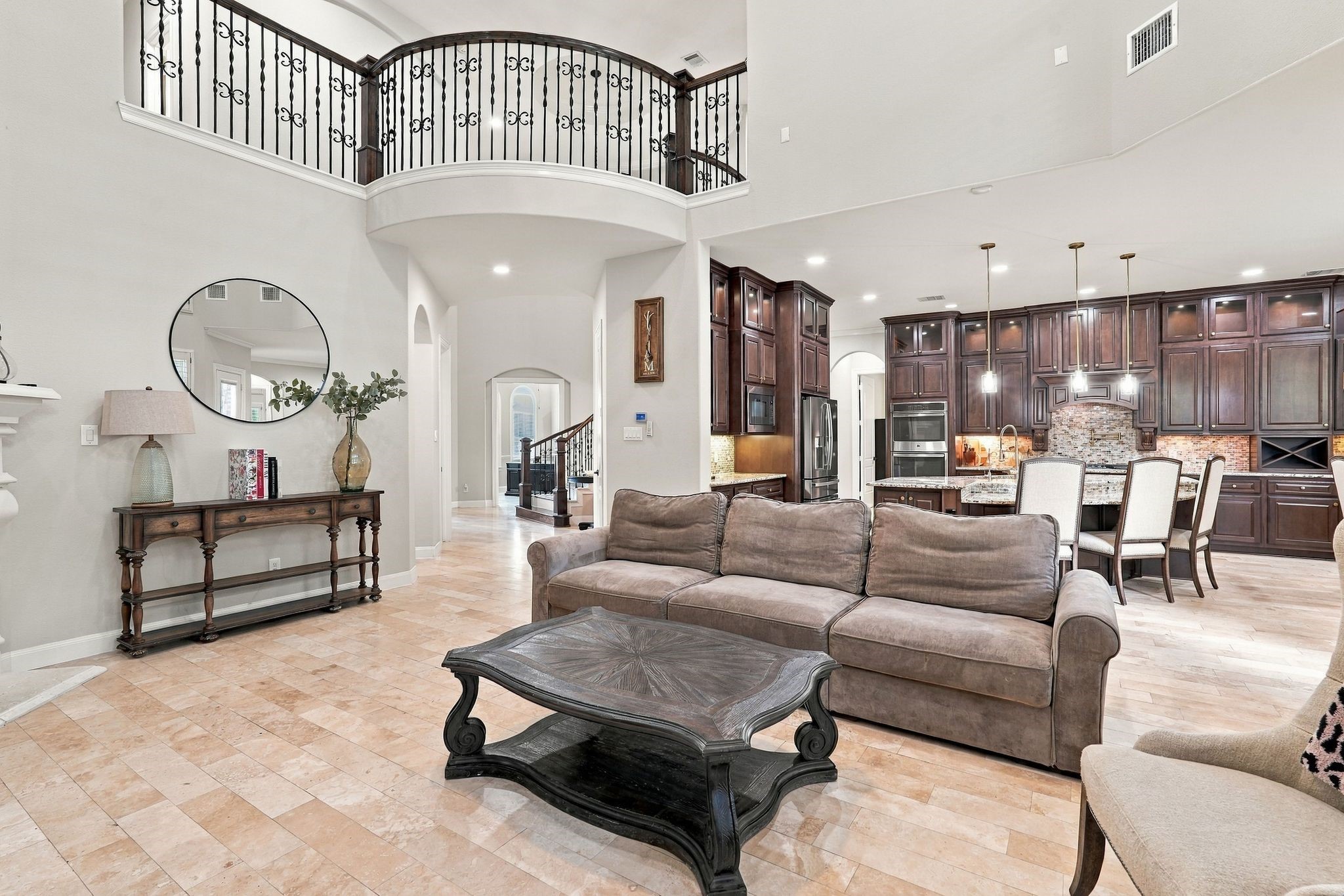 Impressive in both scale and design, this expansive two-story living room showcases soaring ceilings, a dramatic curved second-story balcony with wrought-iron railings, and elegant travertine flooring. The grand, open-concept layout creates an incredible sense of space while flowing seamlessly into the gourmet kitchen, perfect for entertaining on any scale.