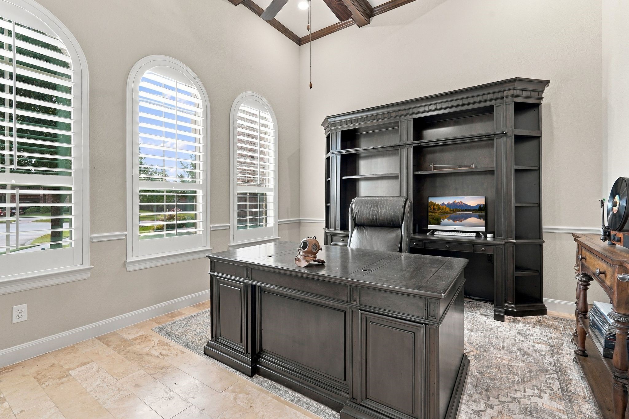 Elegant private home office featuring soaring ceilings with decorative wood beams, three beautiful arched windows with plantation shutters, and natural stone flooring. Abundant natural light creates an inviting and functional workspace, ideal for working from home.