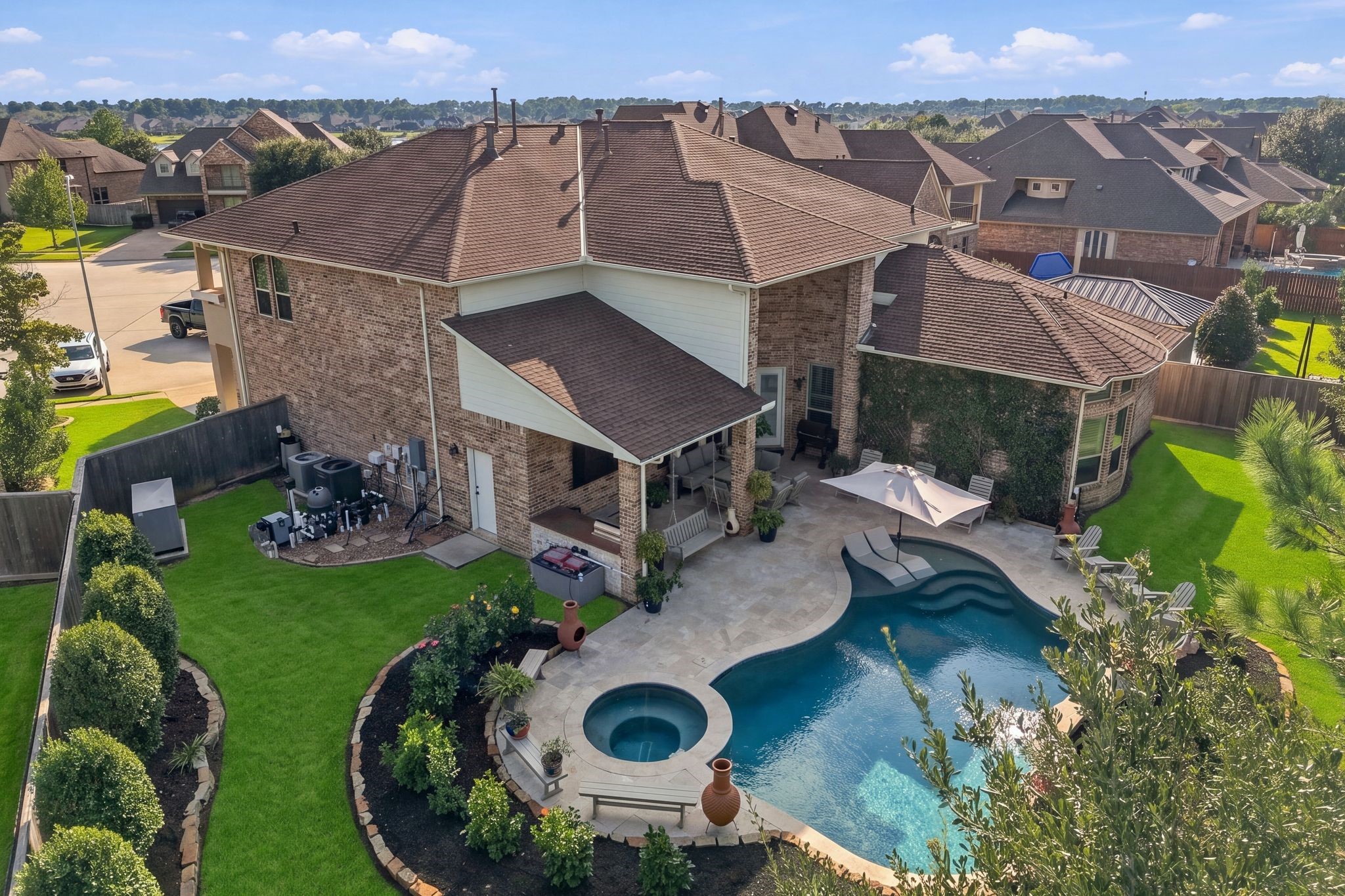 Stunning aerial view of the resort-style backyard showcasing a custom-designed pool and spa, expansive travertine decking, covered outdoor living area with kitchen, and beautifully manicured landscaping. Generous green space and mature trees provide the perfect balance of luxury outdoor entertaining and everyday relaxation.