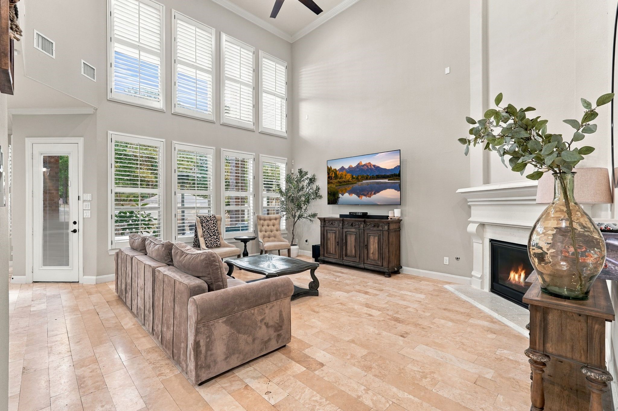 Breathtaking two-story living room showcases soaring ceilings, a dramatic wall of windows adorned with plantation shutters, an elegant fireplace, and beautiful travertine flooring. An abundance of natural light enhances the home's sophisticated architectural design, creating a stunning space for luxurious everyday living and entertaining.