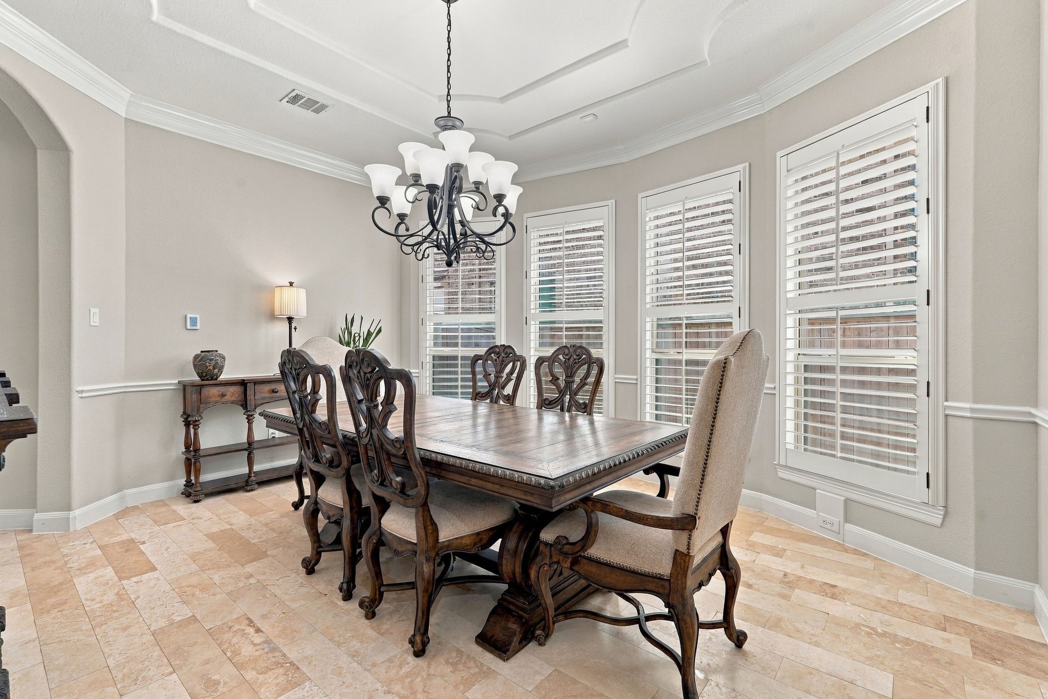 Beautiful formal dining room featuring an elegant tray ceiling, crown molding, chandelier, travertine flooring, and plantation shutters. Large windows provide abundant natural light, creating an inviting space for entertaining and special gatherings.