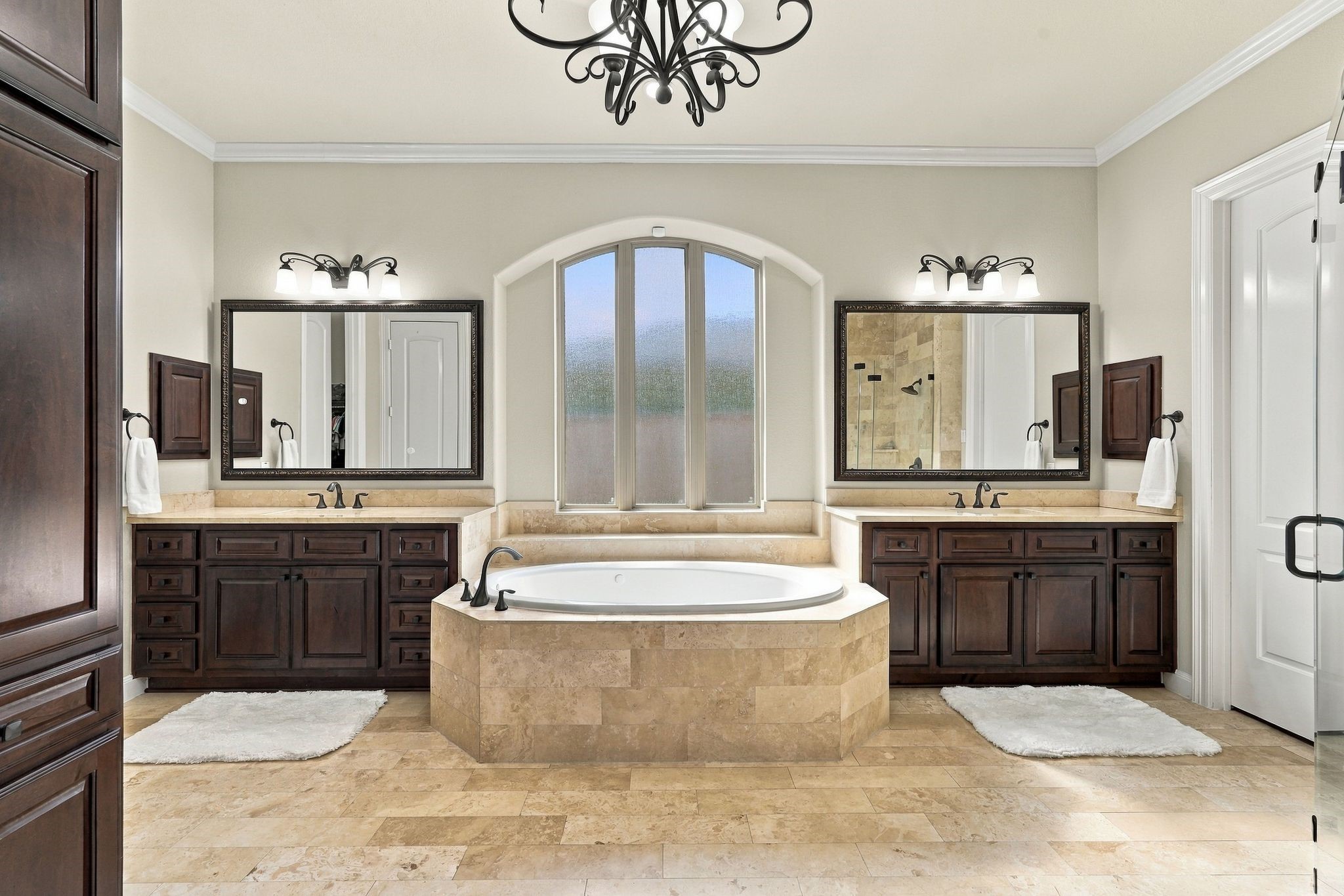 Spa-inspired primary bathroom exudes timeless luxury with a stunning soaking tub framed by an elegant arched window, dual granite-topped vanities with rich wood cabinetry, and beautiful travertine flooring. The spacious, thoughtfully designed retreat offers an exceptional blend of elegance, comfort, and relaxation.