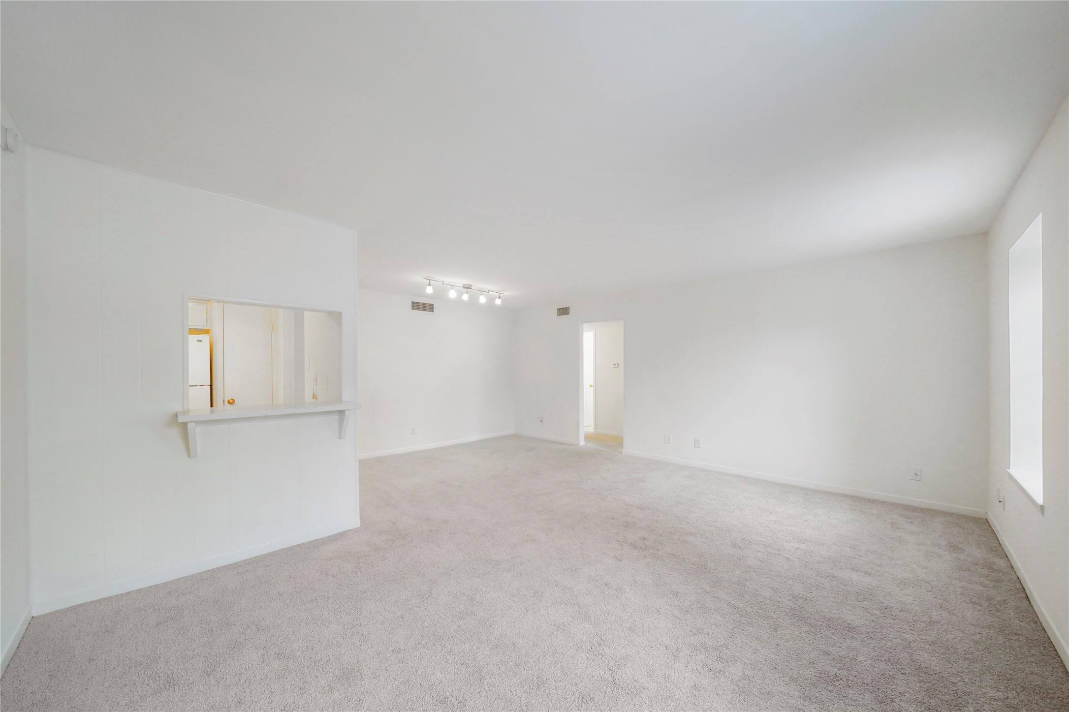Spacious, light-filled living area featuring plush carpet, crisp white walls, track lighting, and a pass-through bar leading to the kitchen.