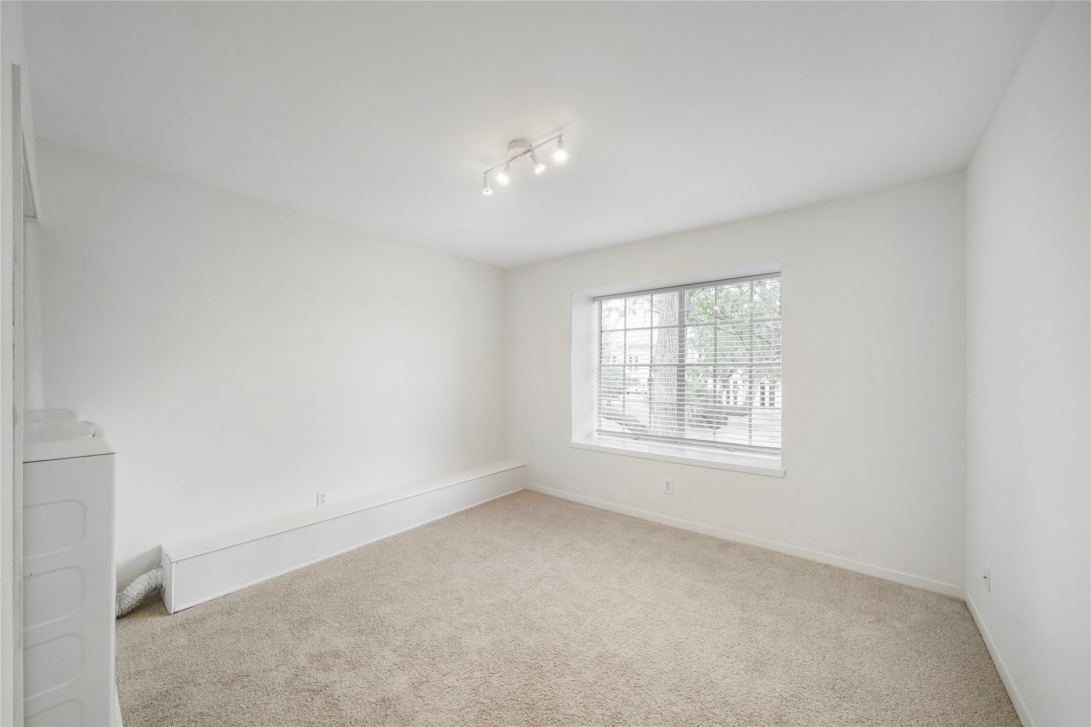 Secondary living space or office area with comfortable carpet, neutral tone walls, track lighting, and a large window offering natural light.