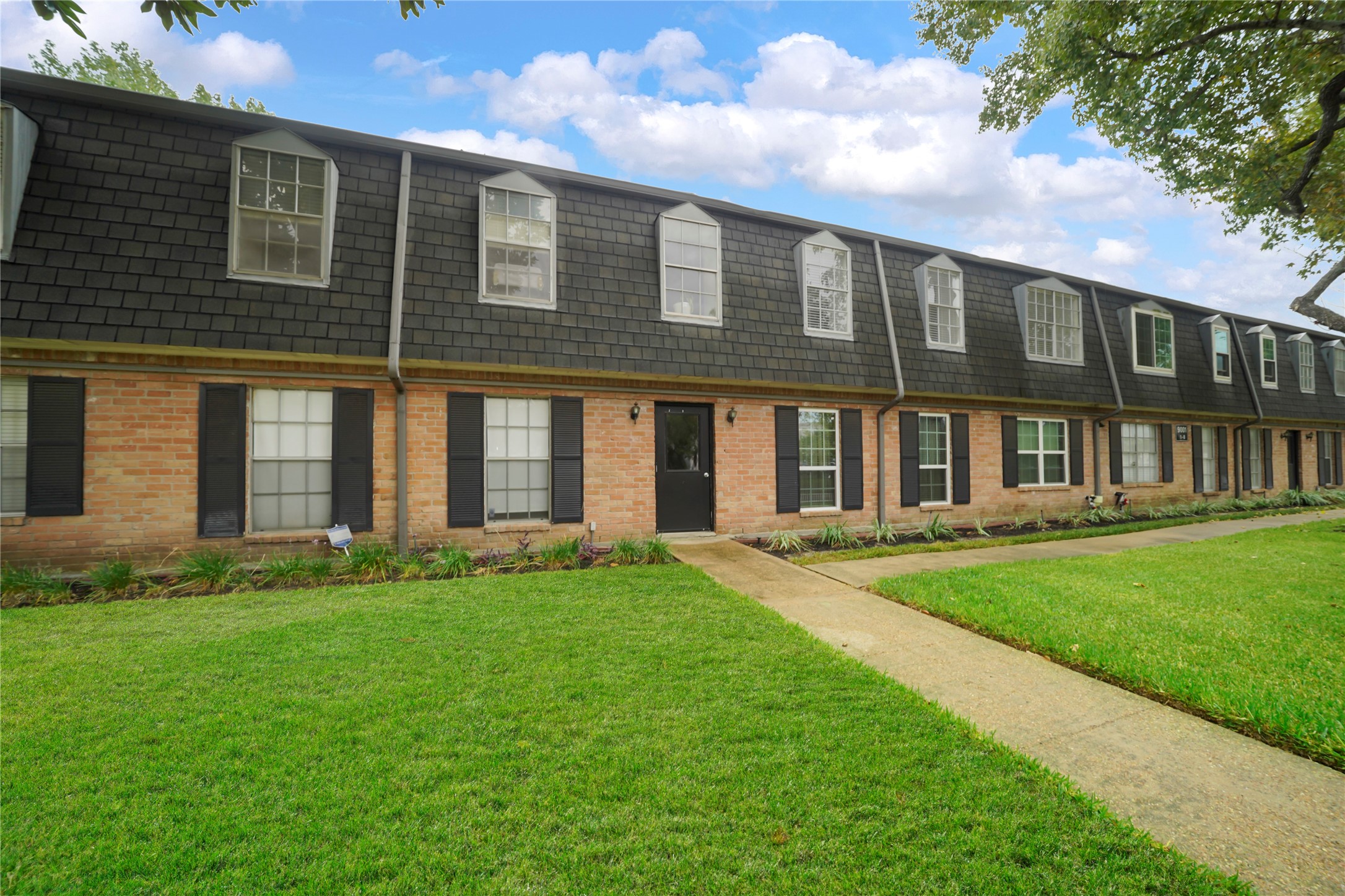 Exterior perspective of the two-story brick and mansard-roof building featuring charming shuttered windows, lush green lawn, and a paved walkway.