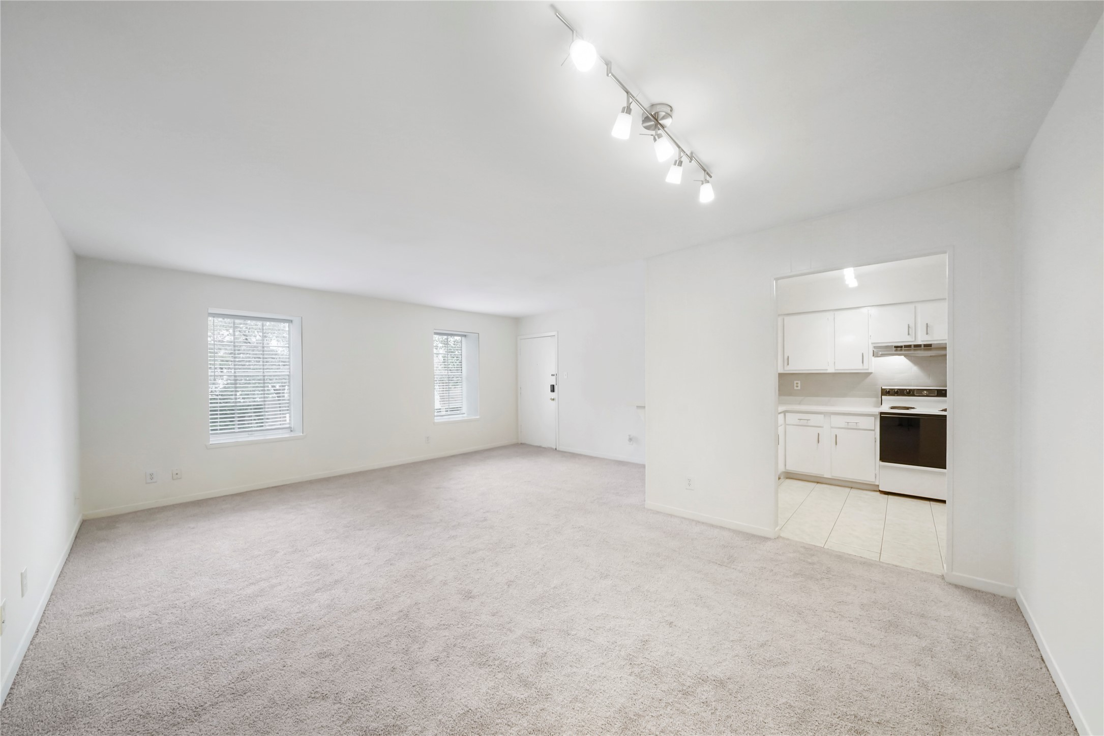 Expansive main living room offering large windows with blinds, plush carpet, track lighting, and direct access to the functional kitchen.