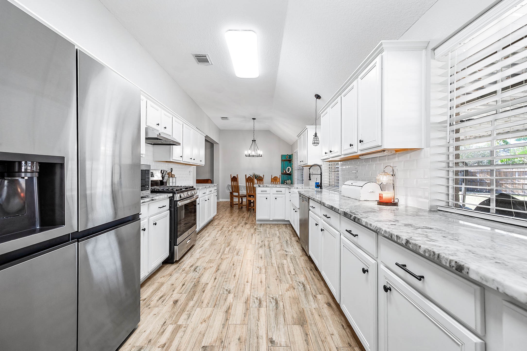 Sleek kitchen featuring modern stainless steel appliances, elegant white cabinetry, and expansive marble countertops, leading to a cozy dining area with abundant natural light.