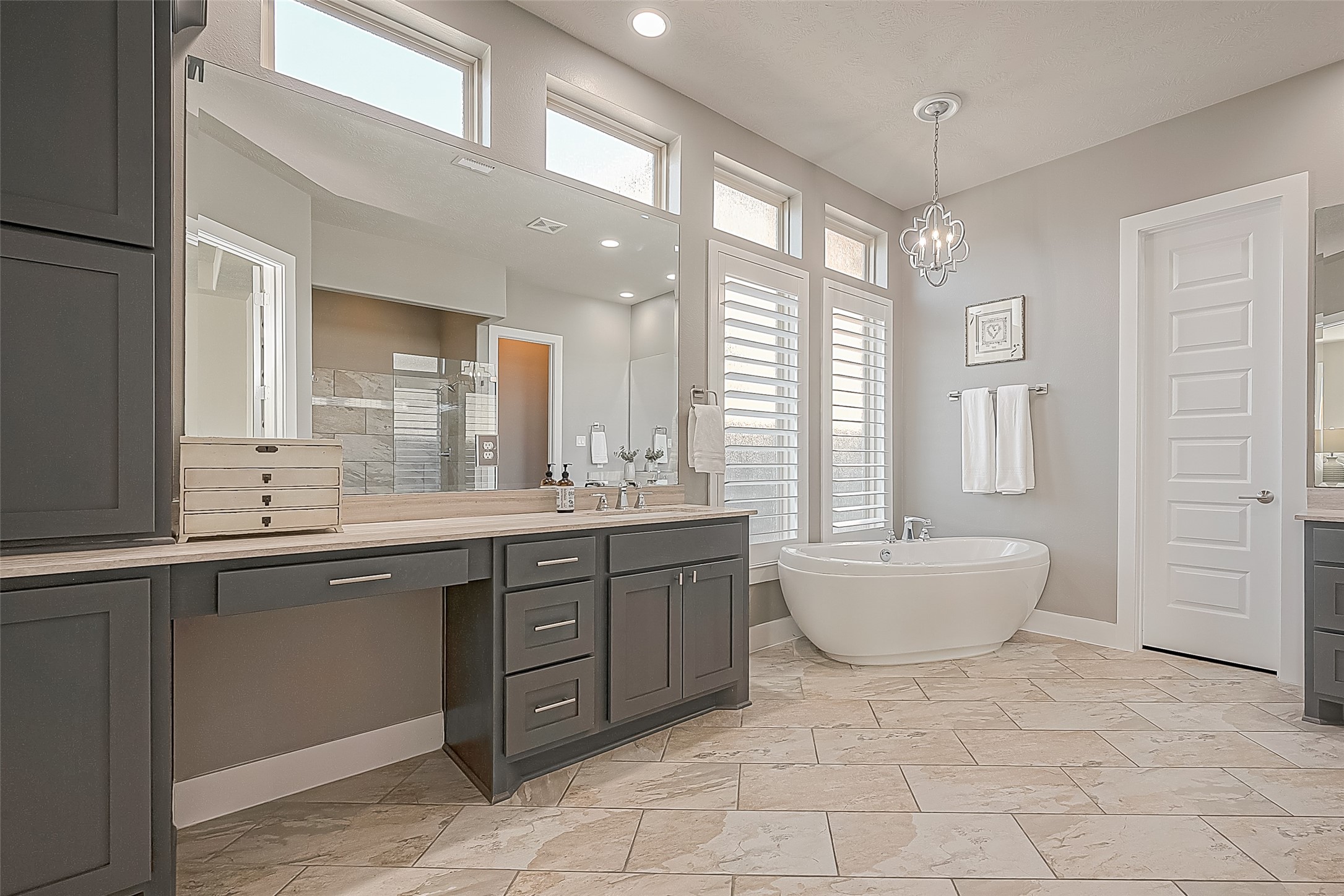 Large primary bath room with large floor tile, Plantation shutters, soaker tub with pendant, dual closets and make-up area on one vanity.