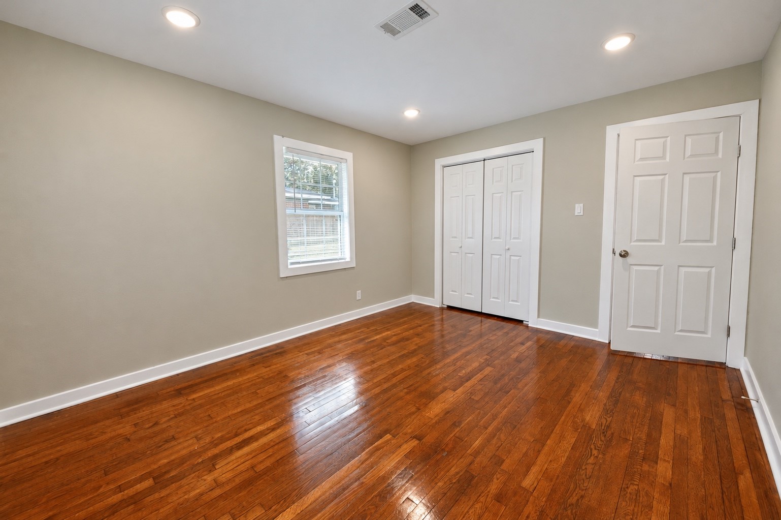 Bright and spacious room featuring polished hardwood floors, neutral walls, and ample natural light from a large window.