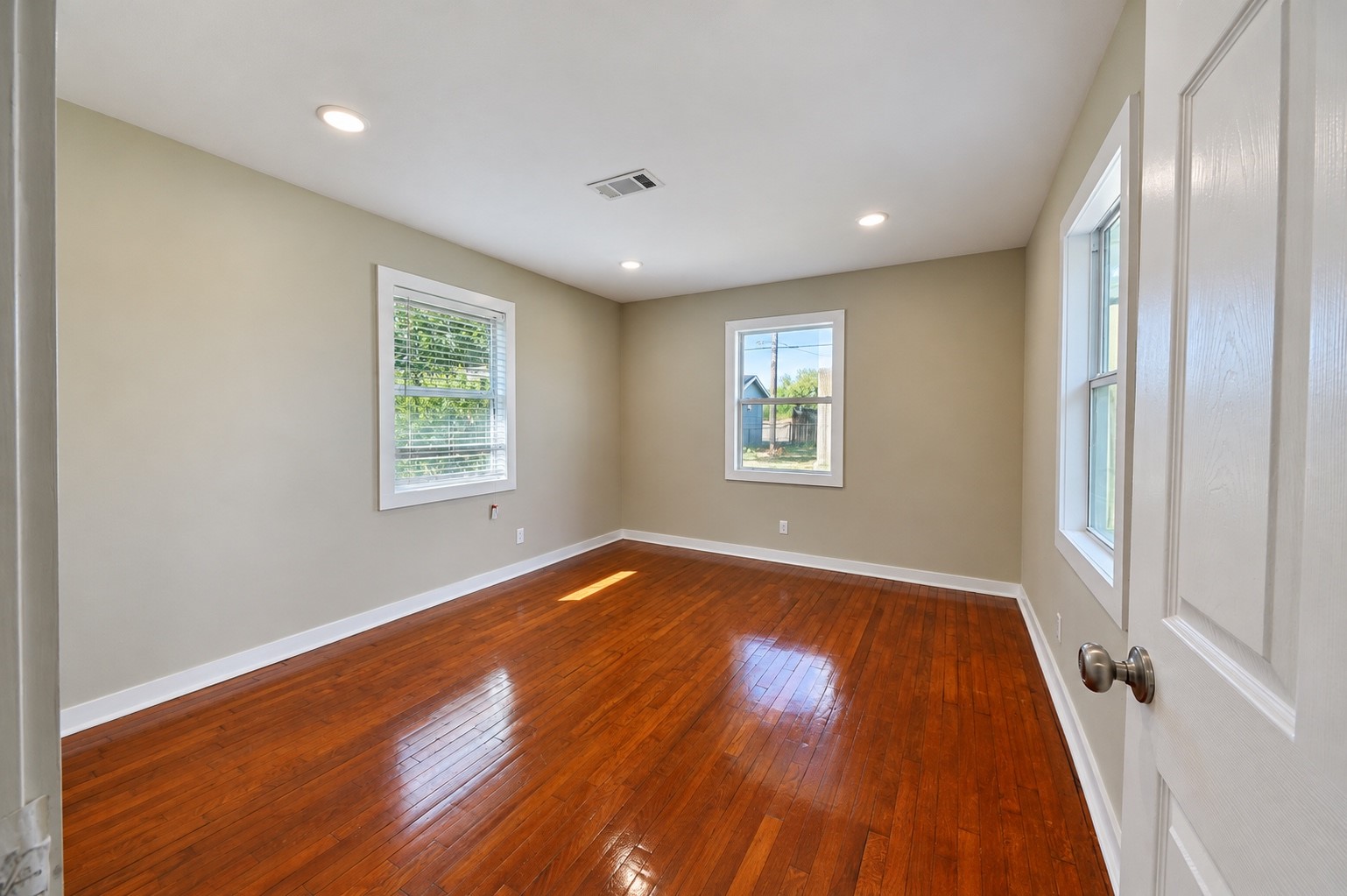 Sunlit room with gleaming hardwood floors, neutral walls, and large windows offering a bright, inviting atmosphere.
