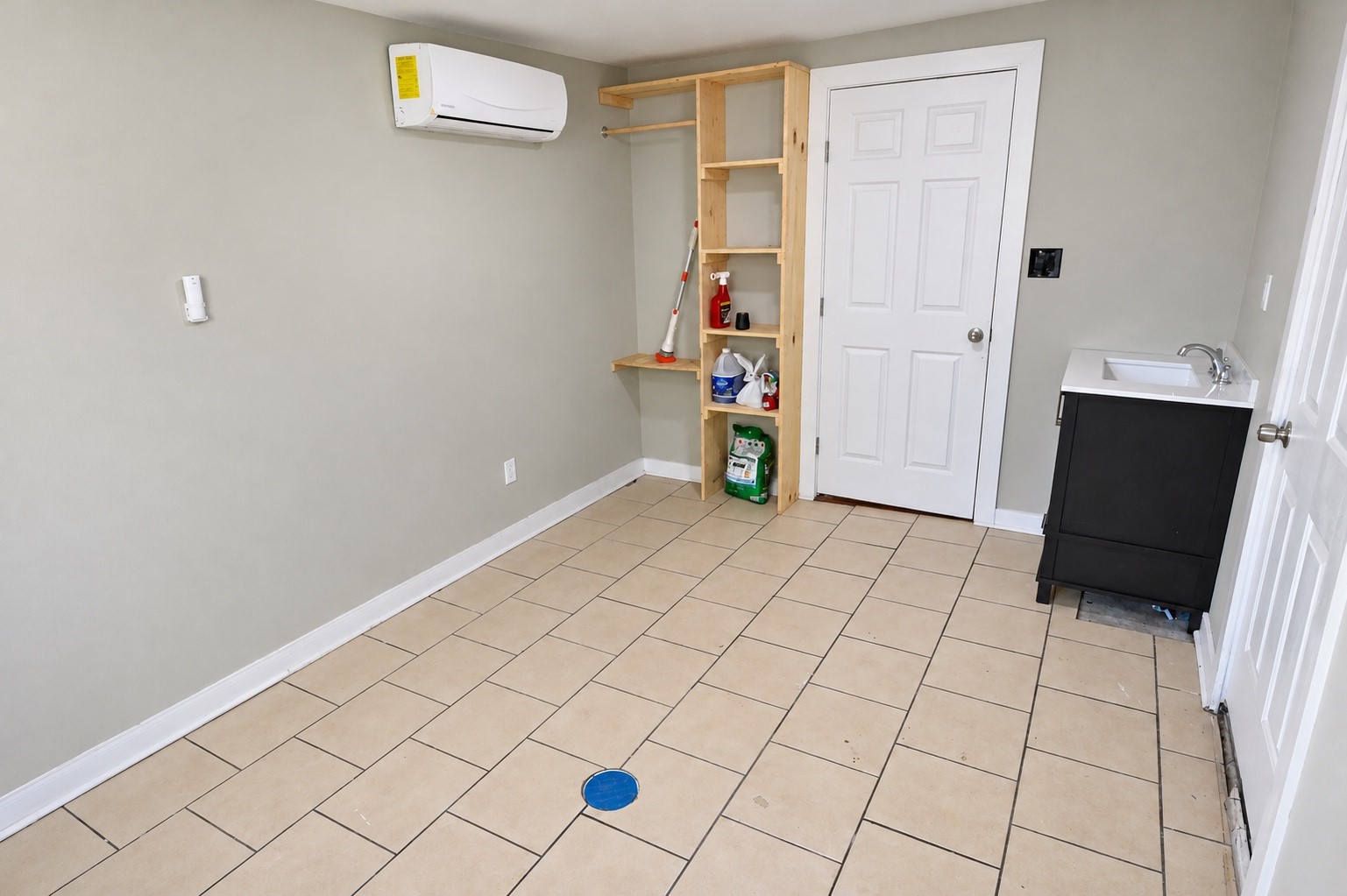 Spacious utility room with tiled flooring, featuring built-in wooden shelving, a convenient sink, and efficient wall-mounted HVAC unit.