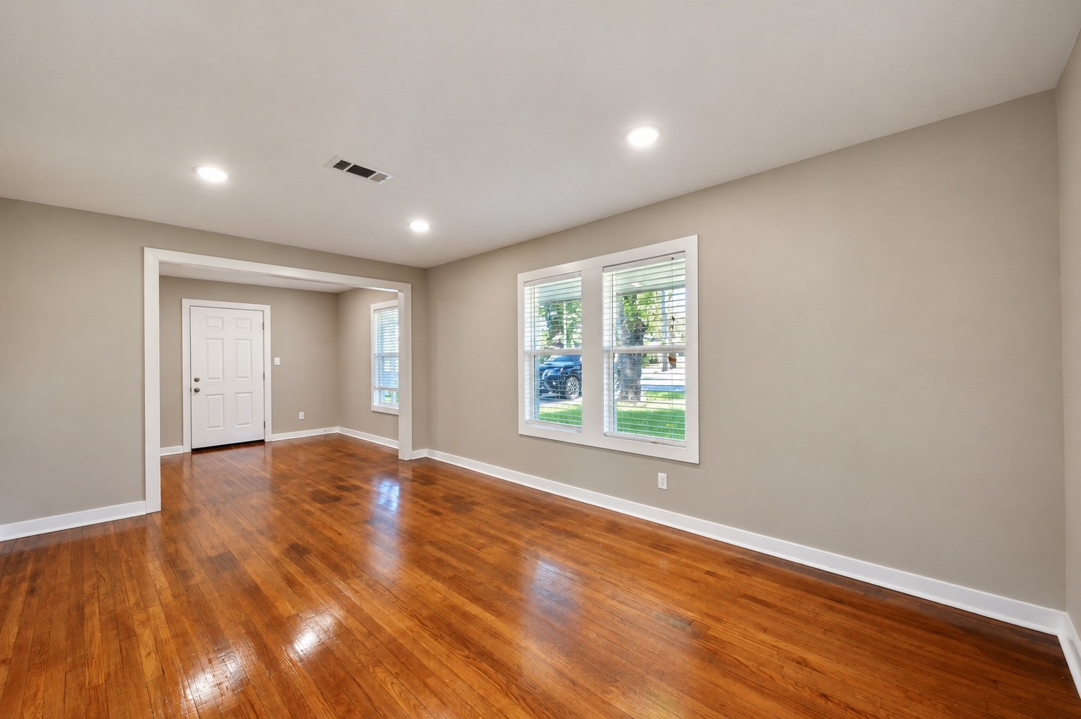 Spacious living area with gleaming hardwood floors, fresh neutral paint, and abundant natural light from large windows, creating a warm and inviting atmosphere.