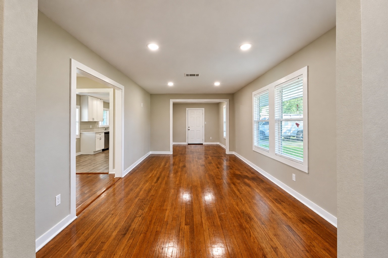 Spacious living area with gleaming hardwood floors, abundant natural light from large windows, and an open layout leading to the kitchen.