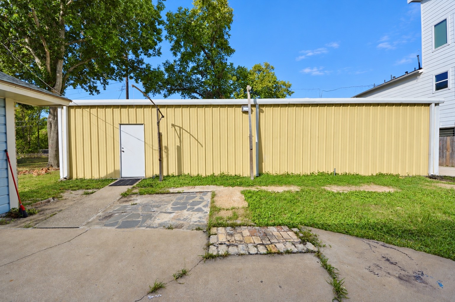 A spacious backyard features a sizable yellow metal storage building with a white door, surrounded by a mix of concrete and grassy areas, ideal for additional storage or a workshop.