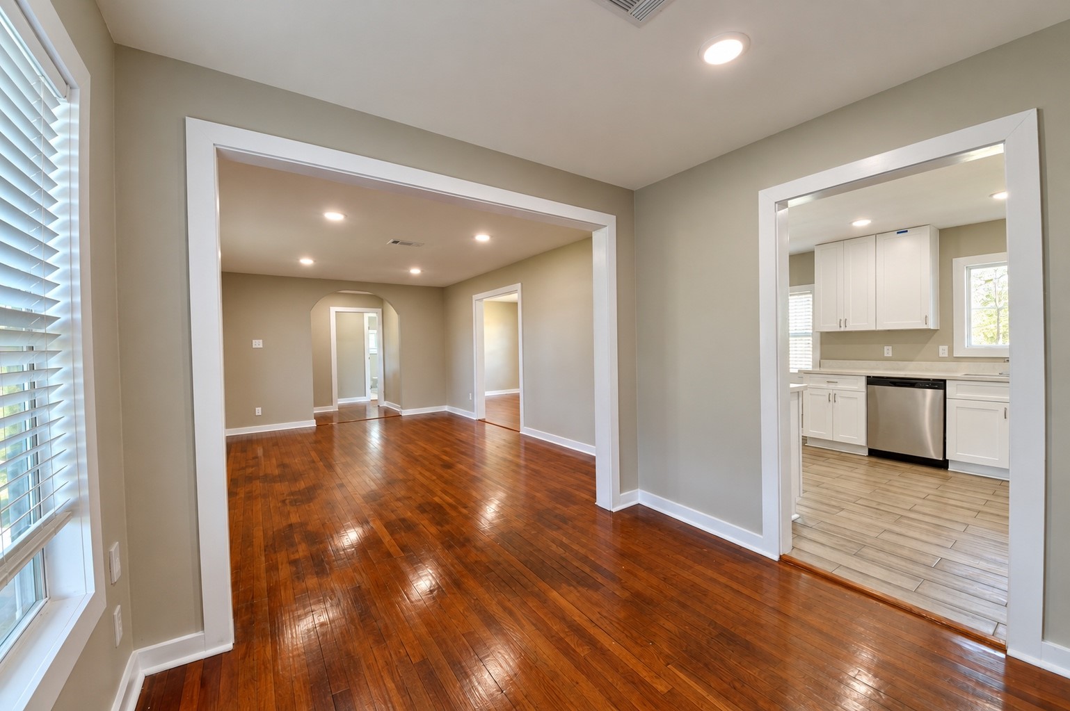 Bright and inviting open-concept living area with rich hardwood floors, recessed lighting, and a seamless flow to the modern kitchen featuring sleek white cabinetry and stainless steel appliances.