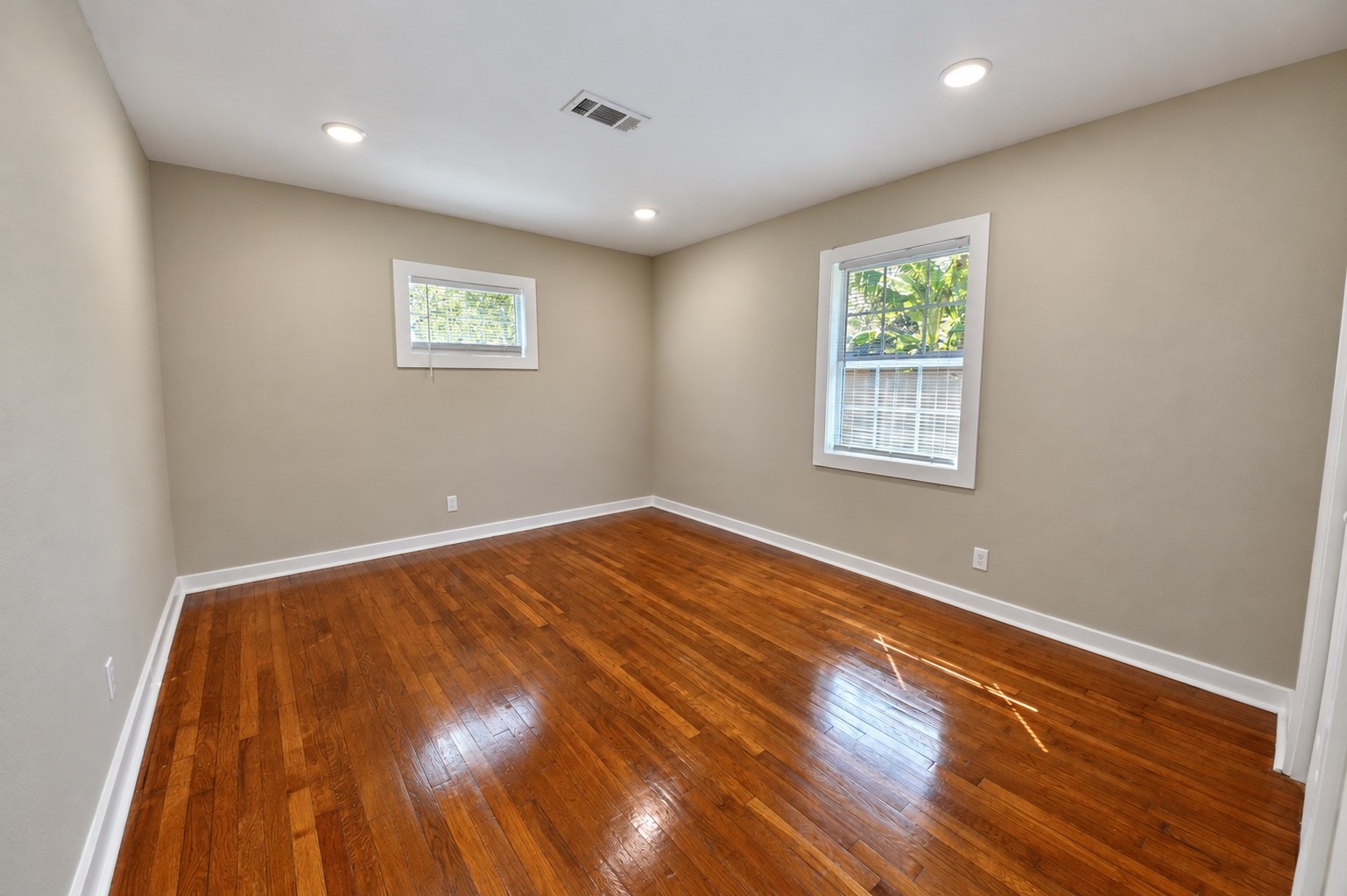 Bright and airy room featuring sleek hardwood floors, neutral walls, and ample natural light from two windows, perfect for a cozy bedroom or home office.