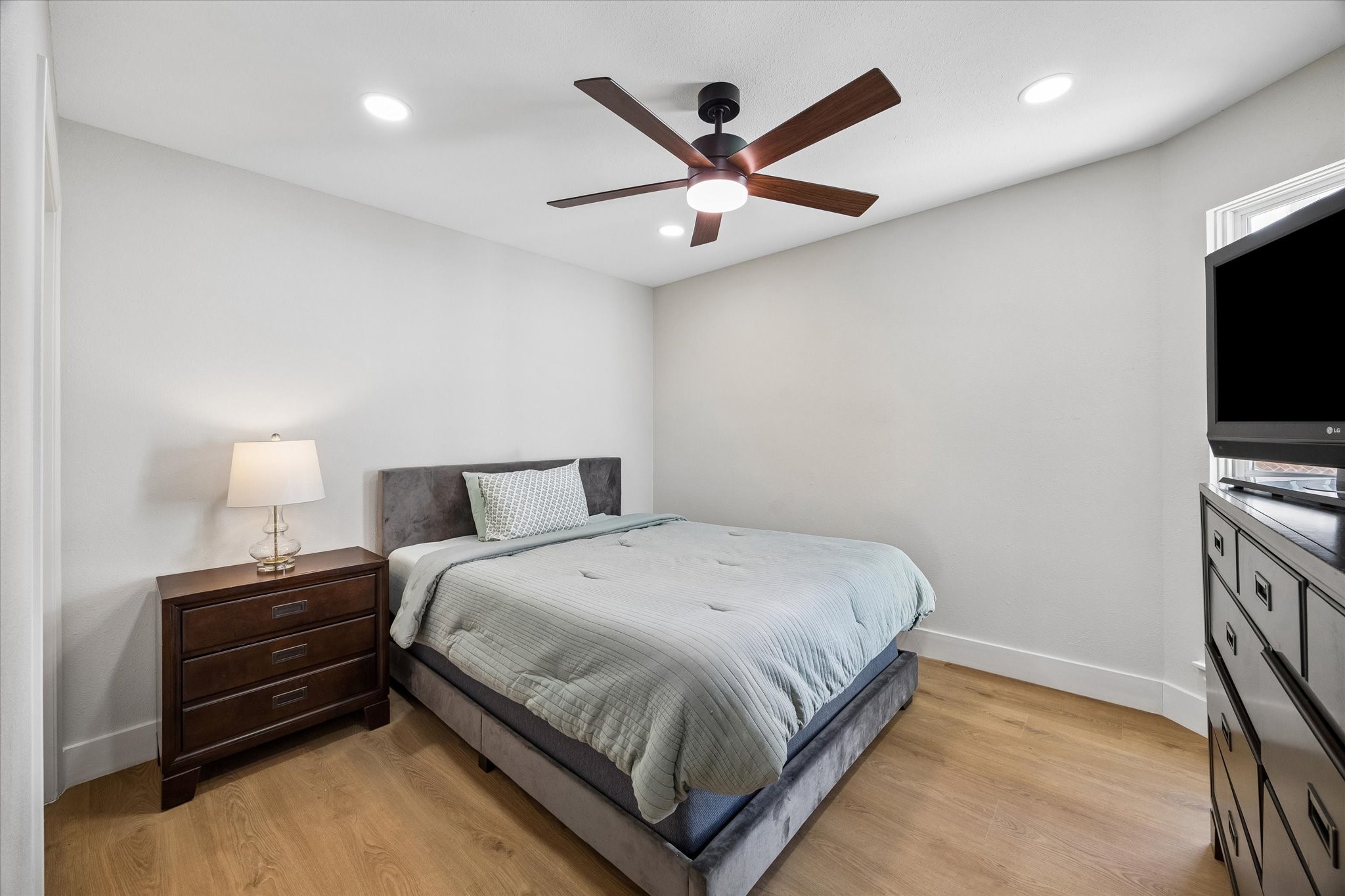 This cozy secondary bedroom features a modern ceiling fan, sleek wood looking floors, and ample natural light from a corner window.