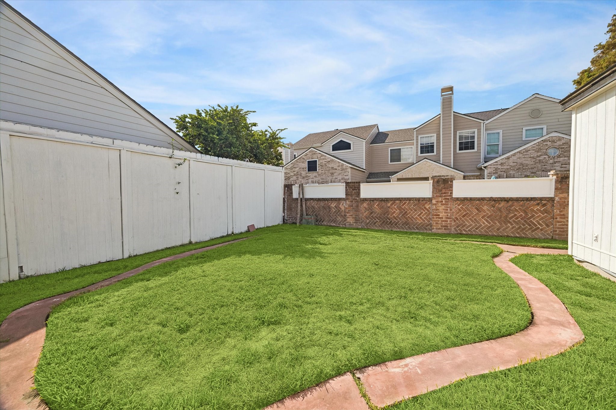 Charming backyard with lush green lawn, bordered by a privacy fence and a unique brick wall, featuring a curved pathway for easy accessibility.