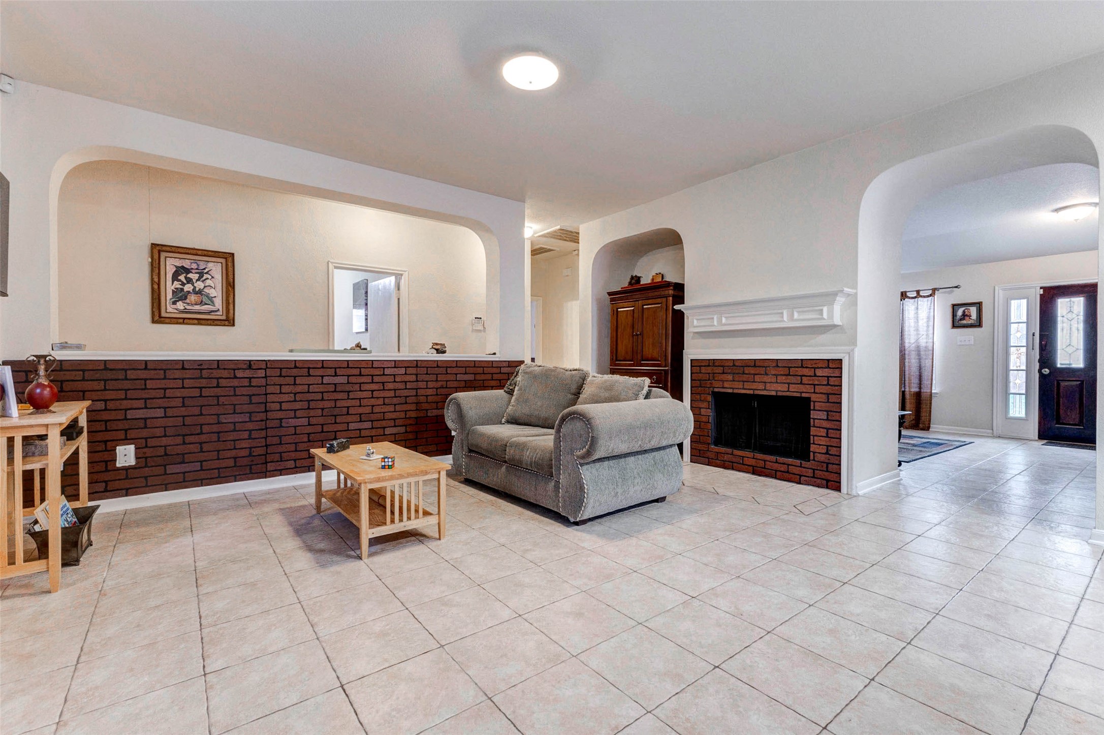 Spacious living area featuring an open layout with durable tile flooring and a cozy brick-accent fireplace that adds warmth and character. Arched openings and a half-wall design create visual separation while maintaining an airy, connected feel. Neutral tones throughout provide a versatile backdrop, while natural light enhances the inviting atmosphere, making this space perfect for relaxing or entertaining guests.