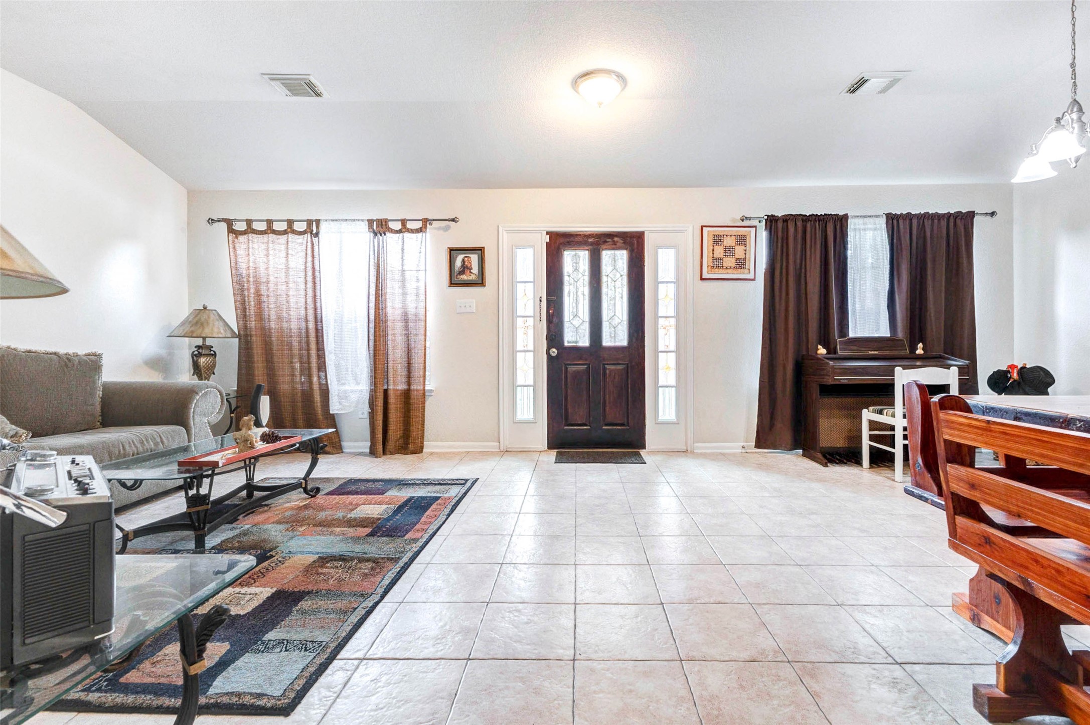 Bright and welcoming entry and living space featuring a wide-open layout with tile flooring and abundant natural light. The front door is framed by sidelights and large windows, creating a warm first impression. The layout offers flexibility for both seating and dining areas, while neutral tones and clean lines provide a timeless backdrop ideal for everyday living and entertaining guests.