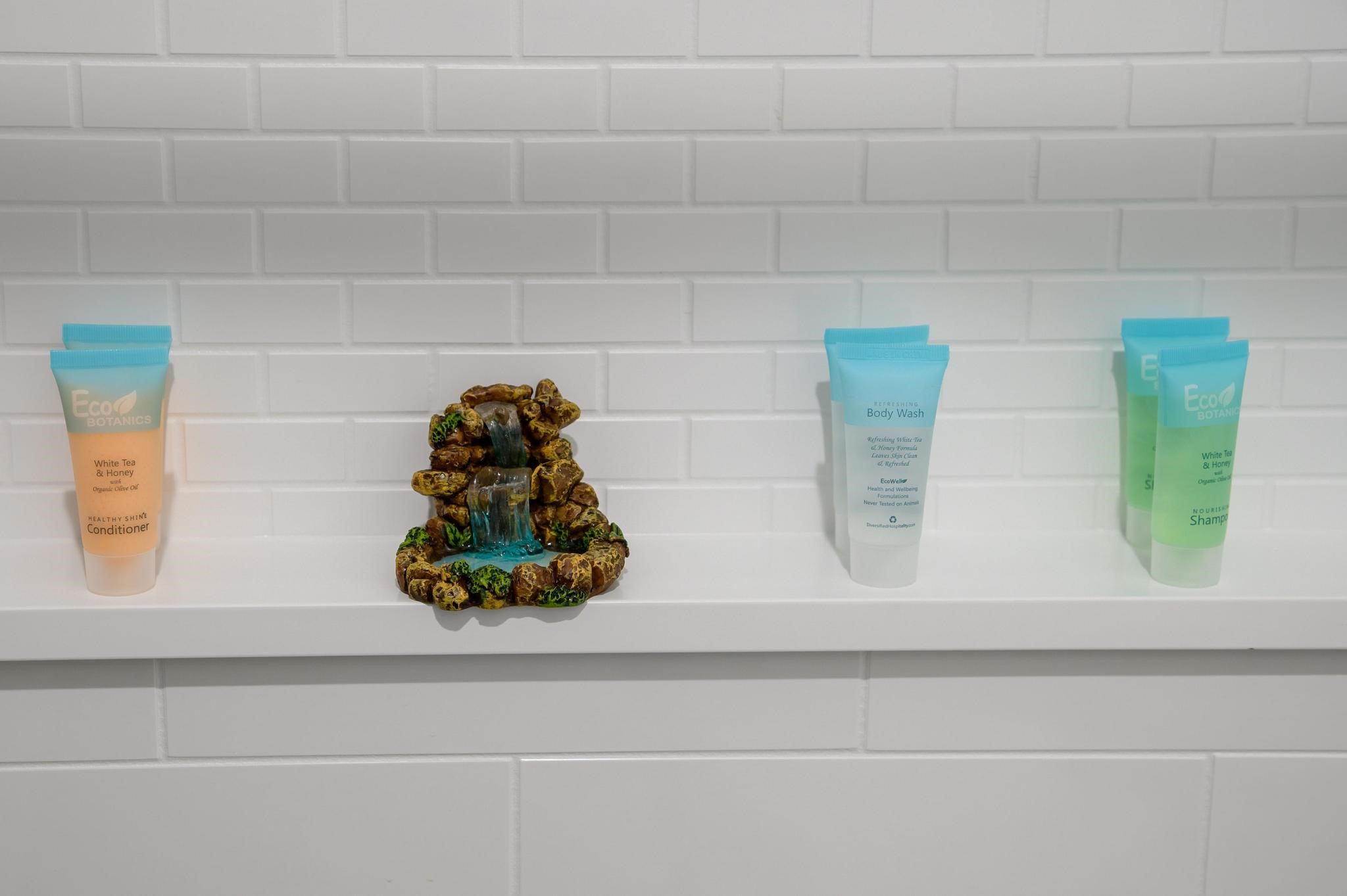 A stylish bathroom shelf features neatly arranged toiletries and a charming miniature waterfall accent against a sleek white subway tile backdrop.