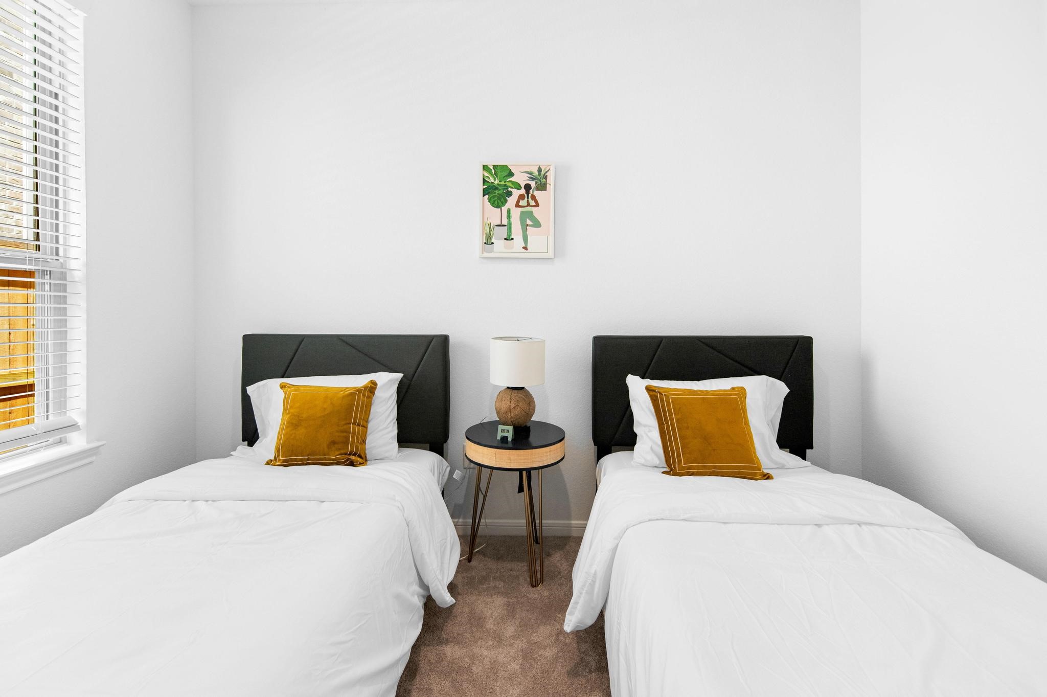 Charming bedroom with two twin beds, each adorned with a mustard accent pillow, flanked by a stylish round nightstand and lamp, all bathed in natural light from a large window.