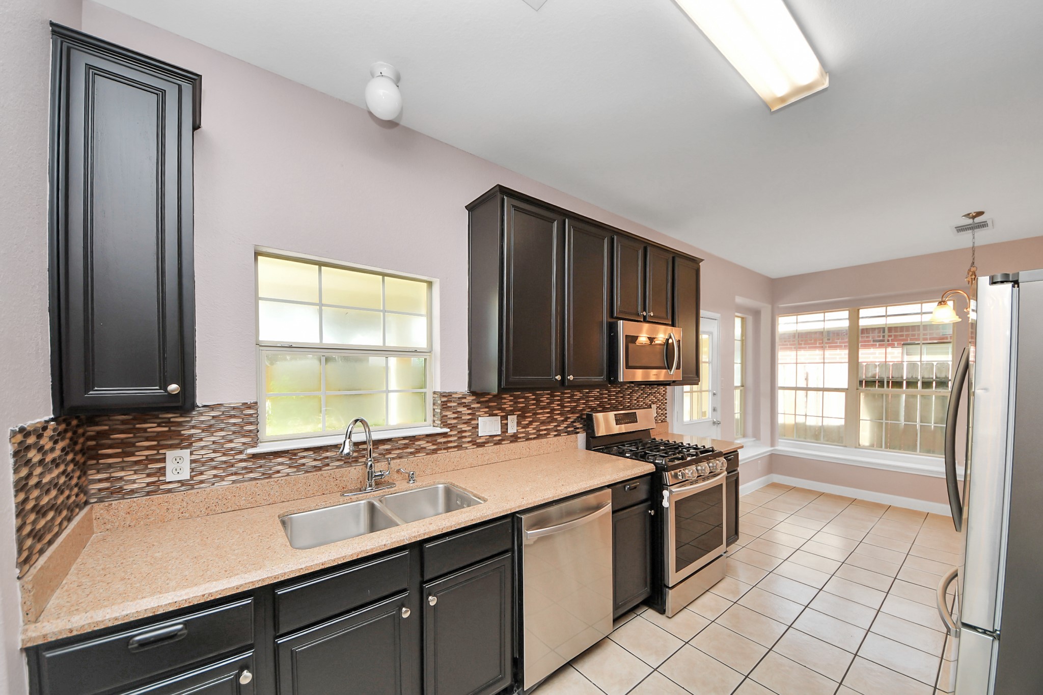 Modern kitchen with sleek dark cabinets, stainless steel appliances, and a stylish backsplash. Ample natural light streams through large windows, highlighting the spacious, tiled floor.