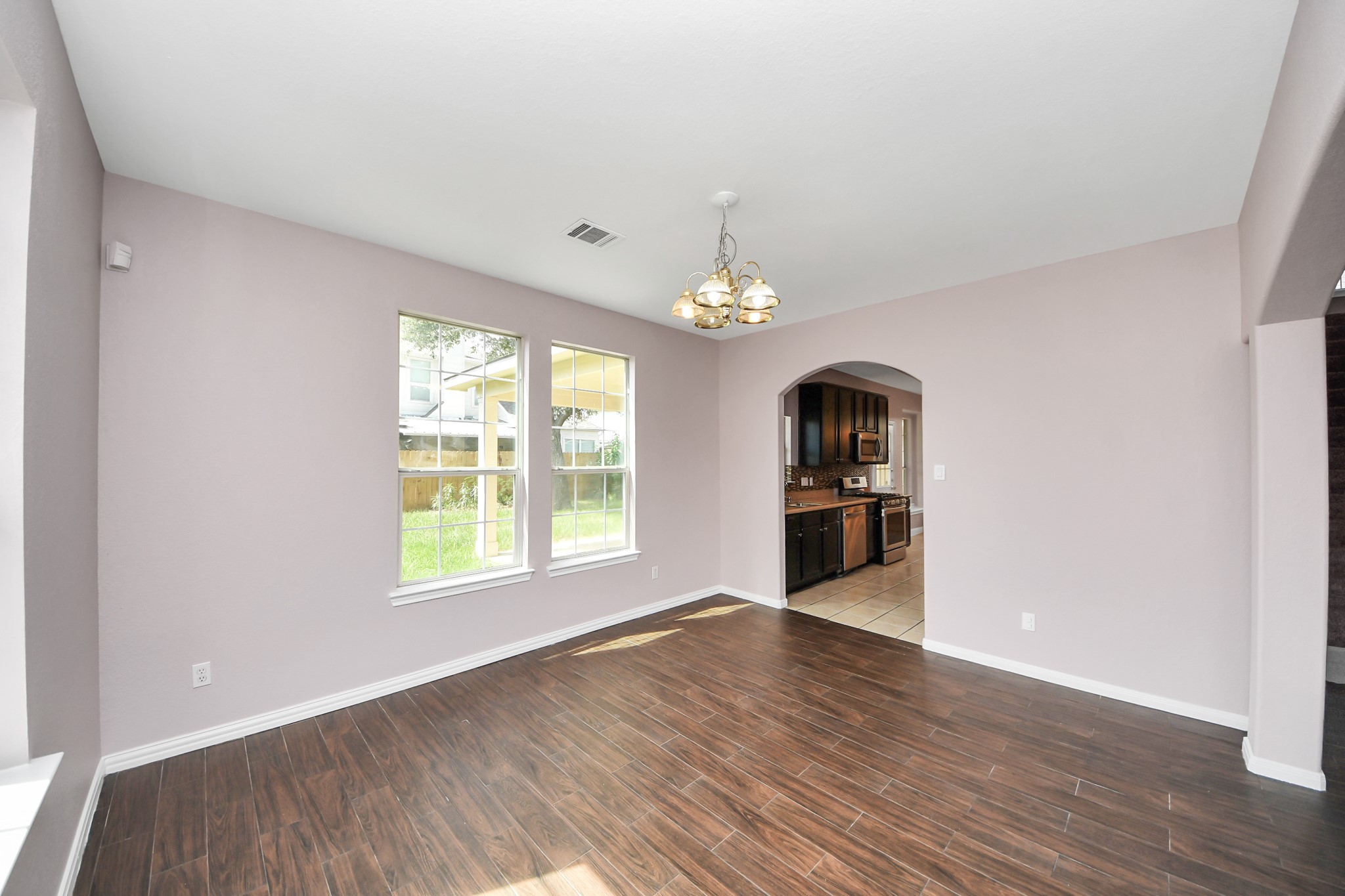 Bright and inviting dining area with elegant wood-style flooring and large windows, seamlessly connected to a modern kitchen through an arched doorway.