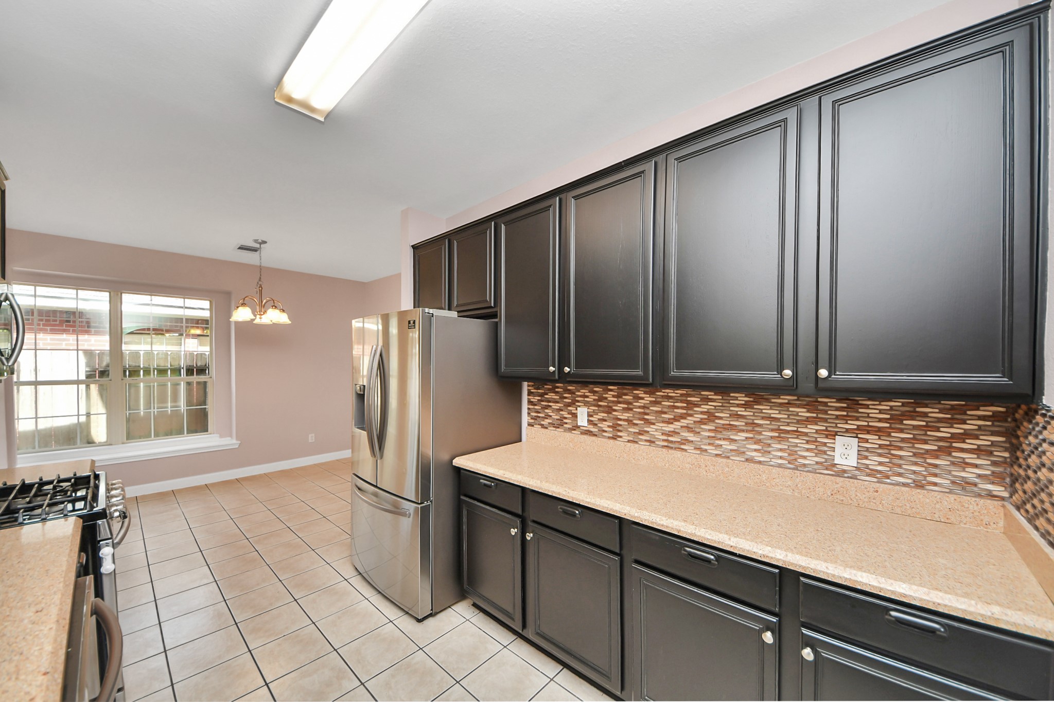 Spacious kitchen with sleek dark cabinetry, modern stainless steel appliances, and a stylish tile backsplash, leading to a bright dining area with large windows.