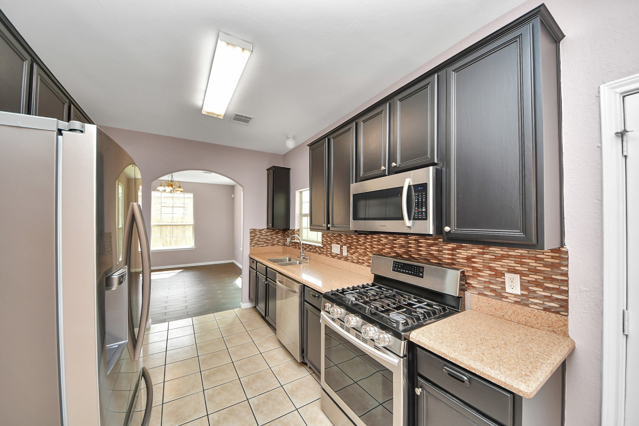 Sleek kitchen featuring dark cabinetry, modern stainless steel appliances, and a stylish backsplash, leading into a bright dining area with large windows.