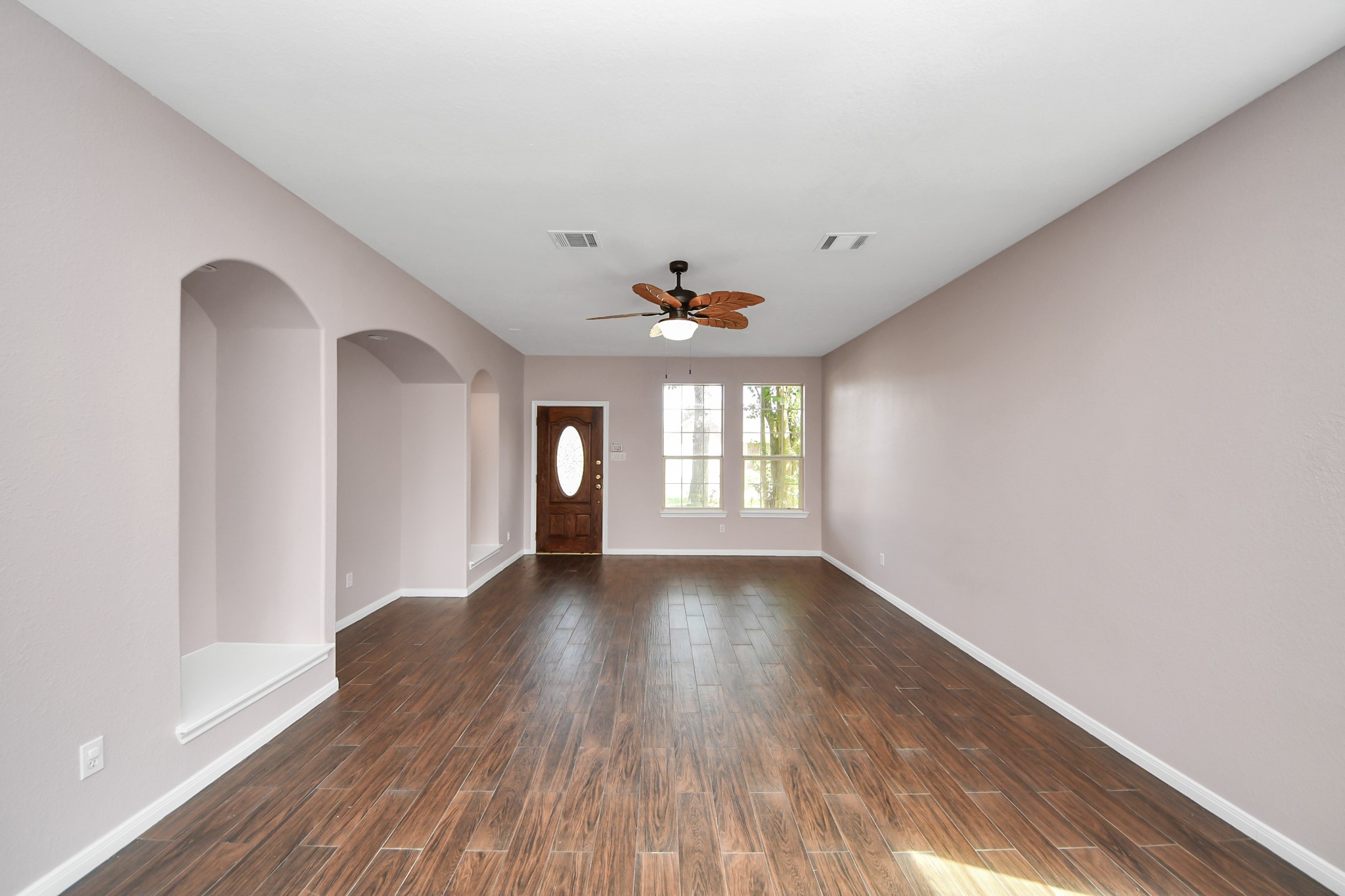 Spacious living room with elegant wood flooring, soft neutral walls, and a stylish ceiling fan, featuring built-in wall niches and ample natural light from large windows.
