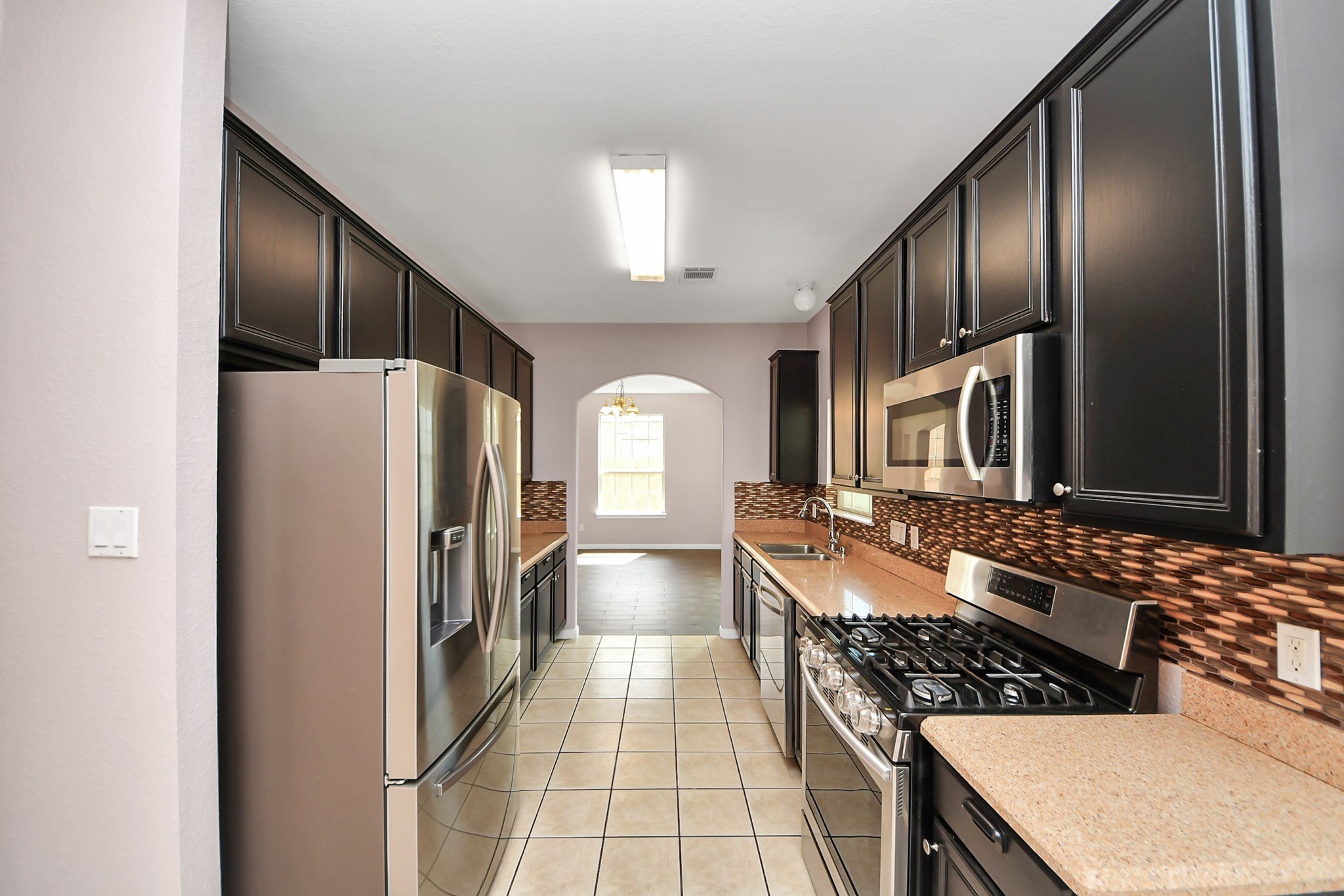 Charming galley kitchen featuring sleek dark cabinetry, modern stainless steel appliances, and a stylish tile backsplash, leading to a cozy dining area filled with natural light.