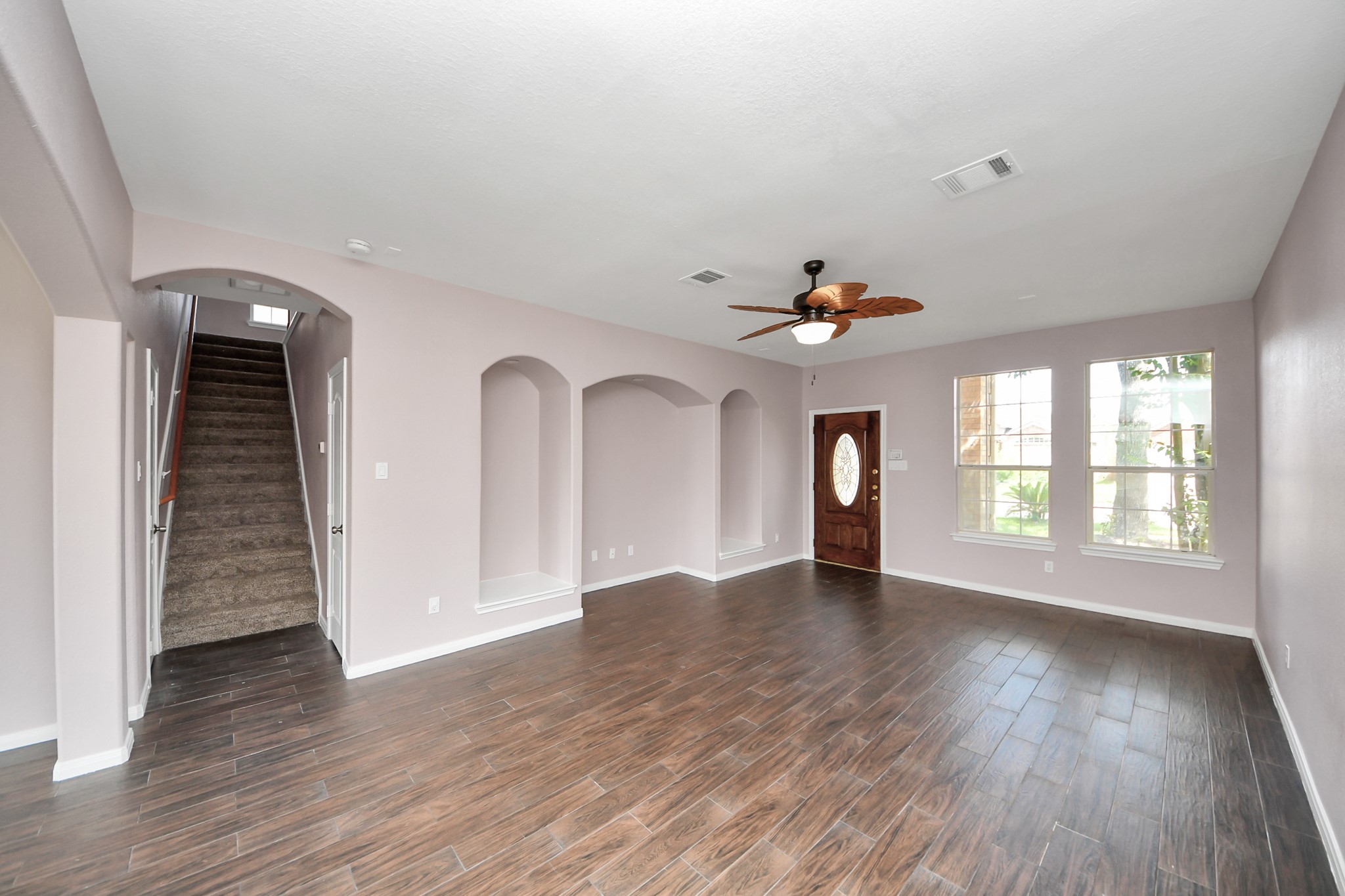 Spacious living room featuring elegant wood-look flooring, recessed niches, and abundant natural light from large windows, complemented by a stylish ceiling fan and an inviting entryway.