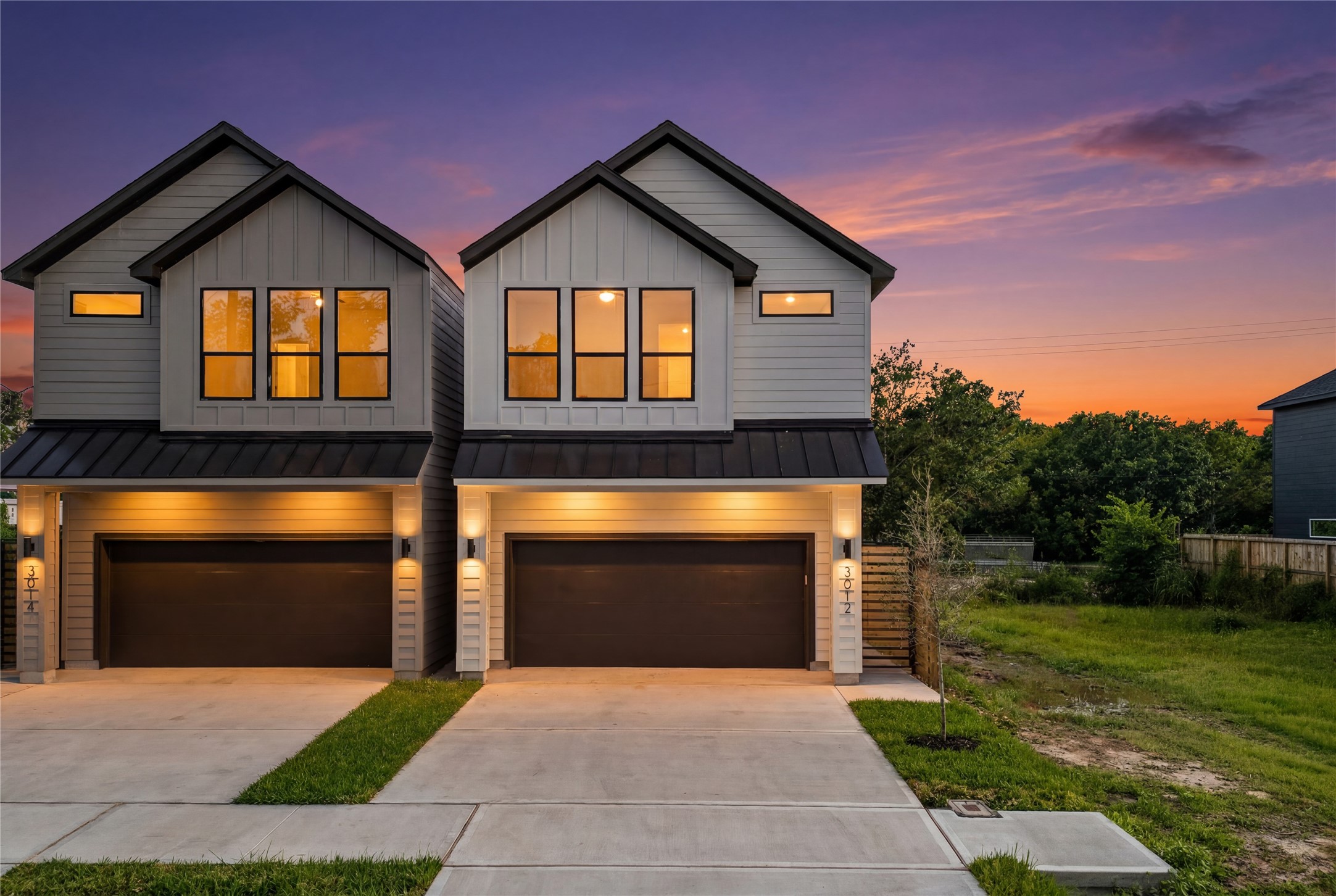 This close-up twilight view focuses on the sleek entryways and dark garage doors. Warm accent lighting glows from the large windows, highlighting the clean lines and modern design elements of the home's exterior.