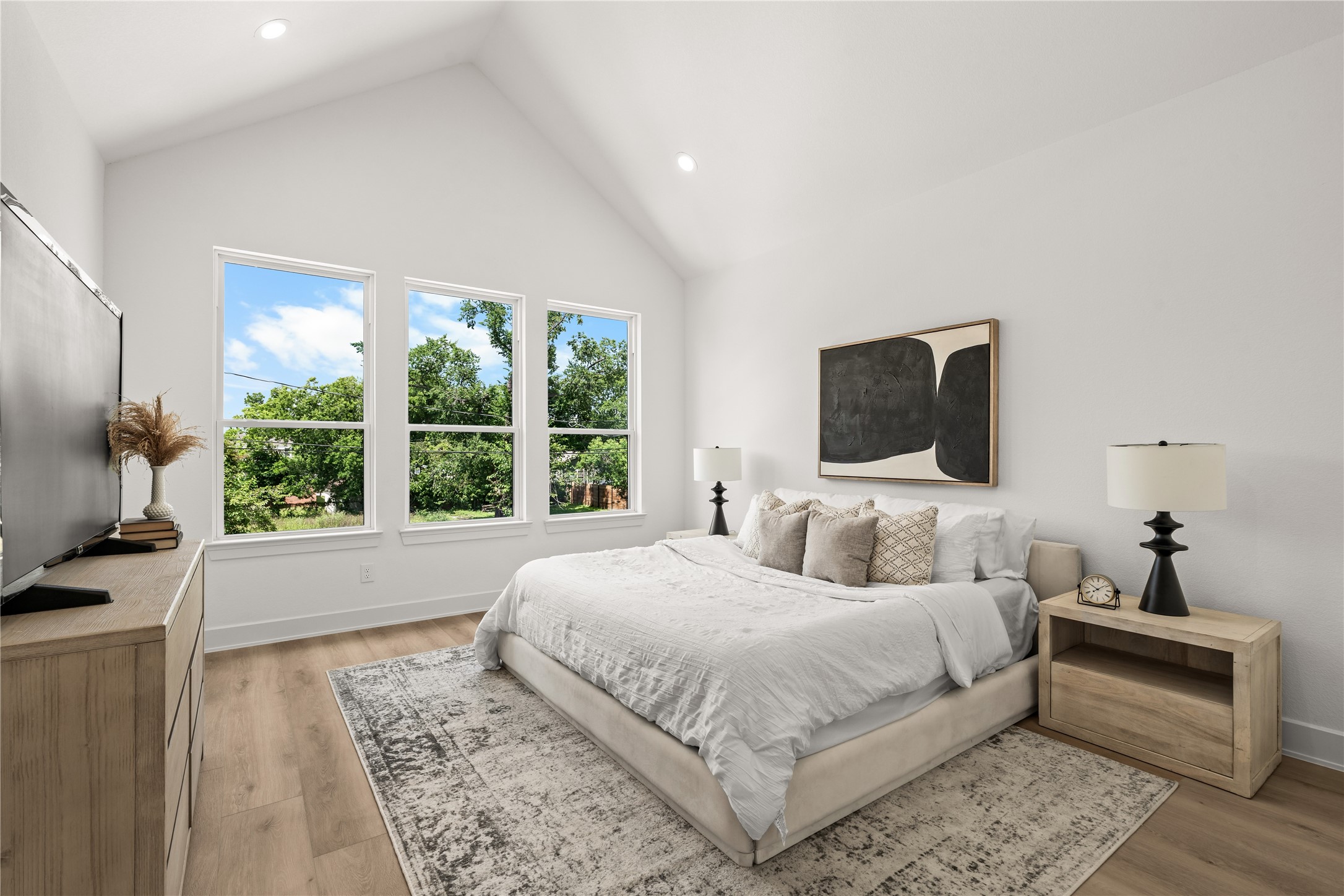 The primary bedroom features a dramatic vaulted ceiling and three large vertical windows that look out onto mature trees. The spacious layout easily fits a king-sized bed, matching nightstands, and a large dresser with room to spare.