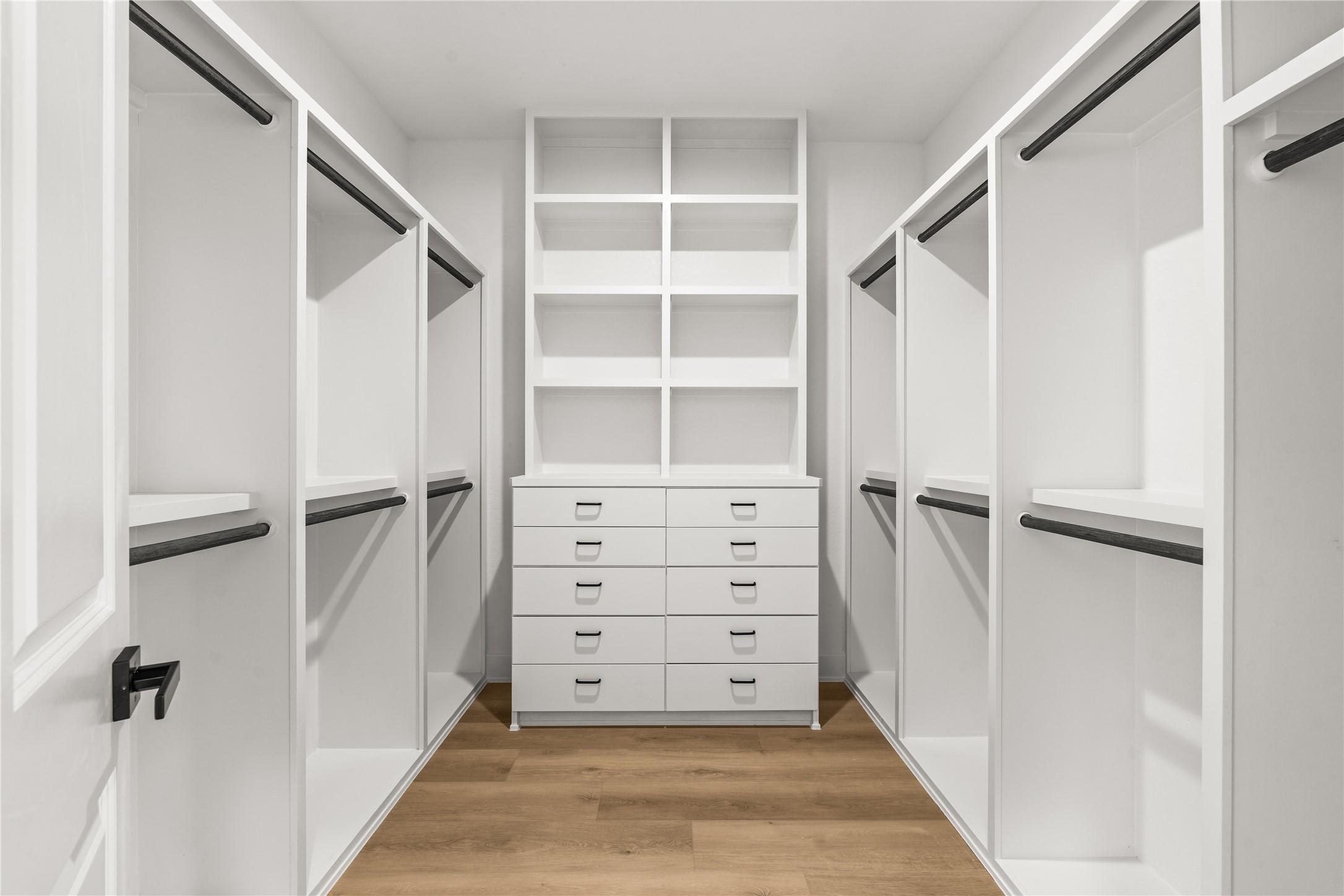 The massive primary walk-in closet features a white custom storage system. A central built-in dresser offers ten drawers with display shelving above, flanked by multiple wardrobe sections equipped with sturdy black hanging rods.