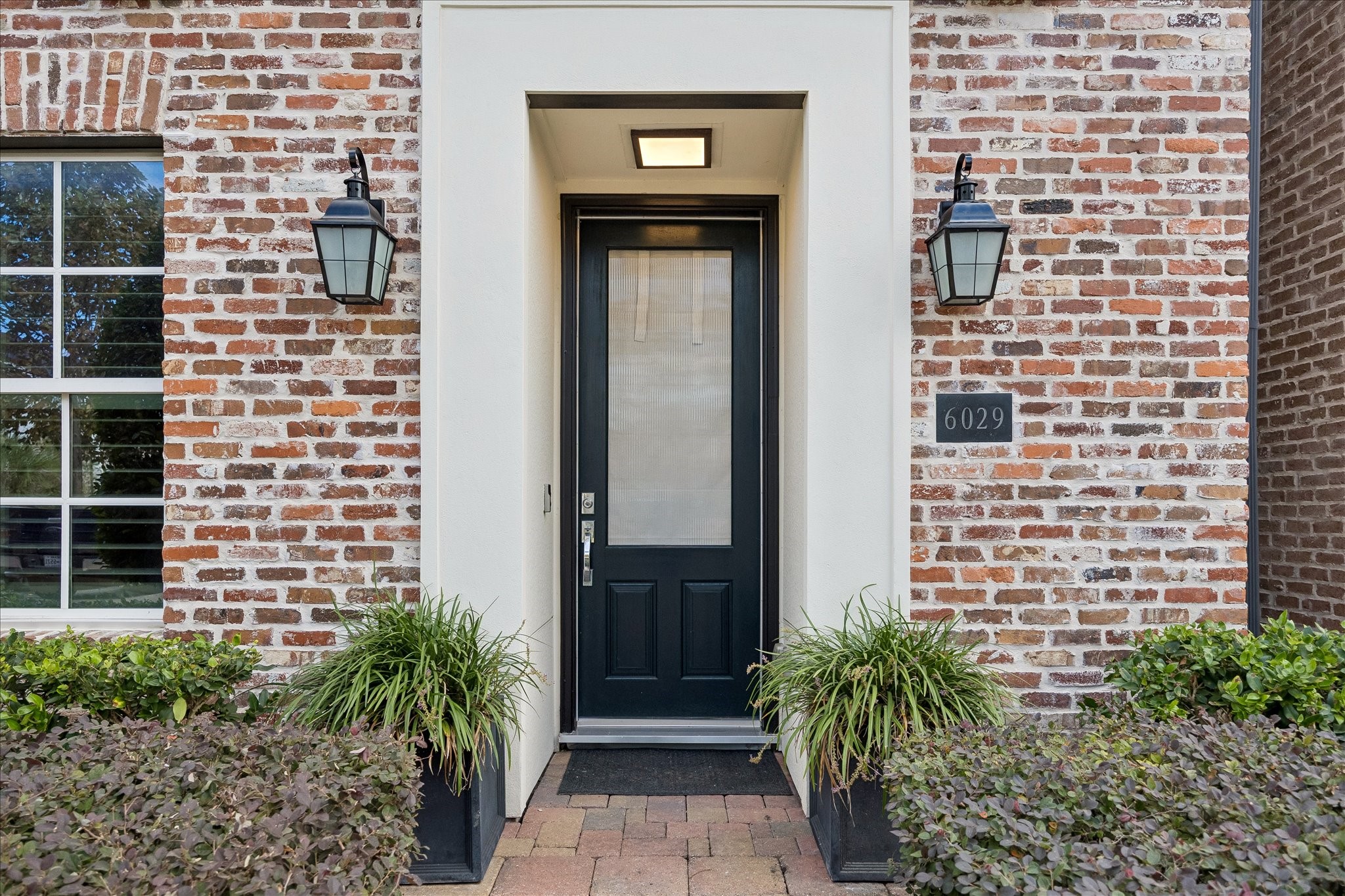 Charming brick exterior with a classic black door and elegant lantern-style lighting, framed by lush greenery for a welcoming entrance.