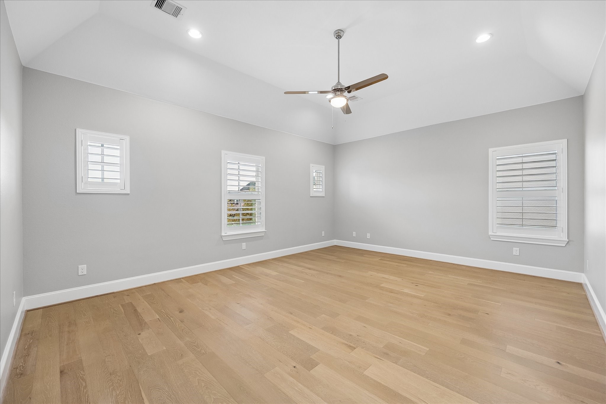 Primary bedroom. Spacious room with high ceilings, elegant light wood flooring, and ample natural light from multiple Plantation shuttered window treatments, complemented by a modern ceiling fan.