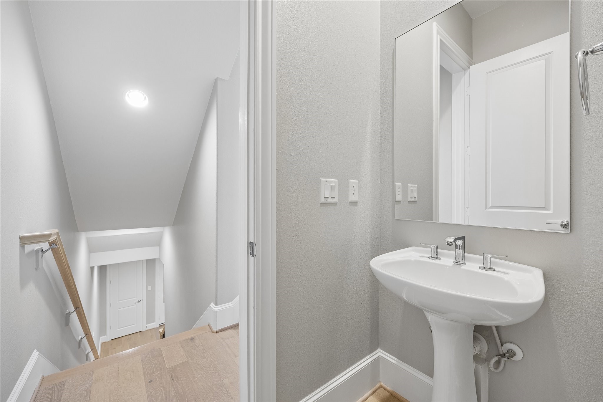 Stylish half-bath featuring a modern pedestal sink and mirror, conveniently located away from the living area and near a bright staircase with sleek wooden handrails.