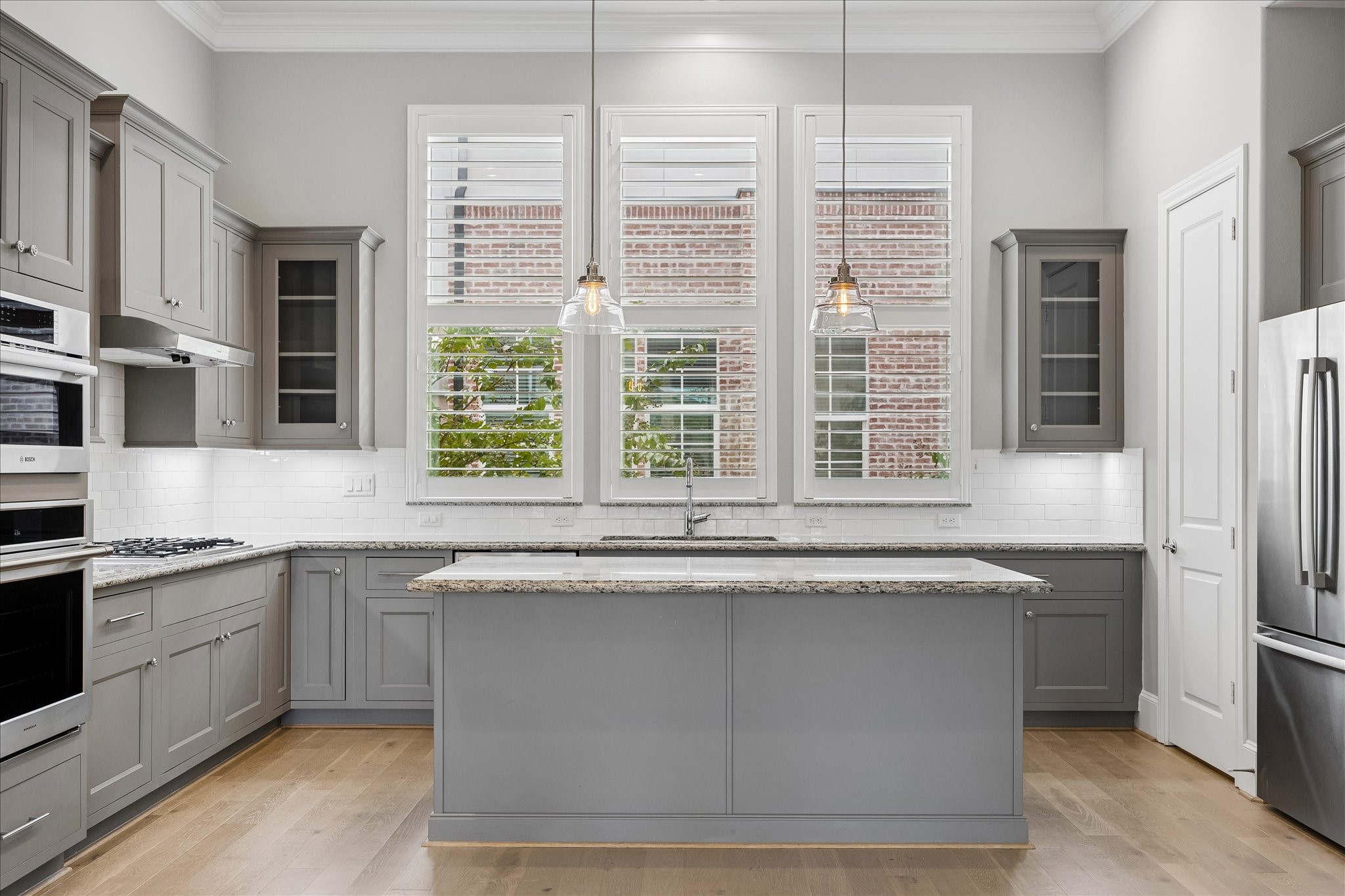 A modern kitchen featuring sleek gray cabinetry, stainless steel appliances, and a spacious island with pendant lighting, complemented by large windows with Plantation Shutters that offer ample natural light.