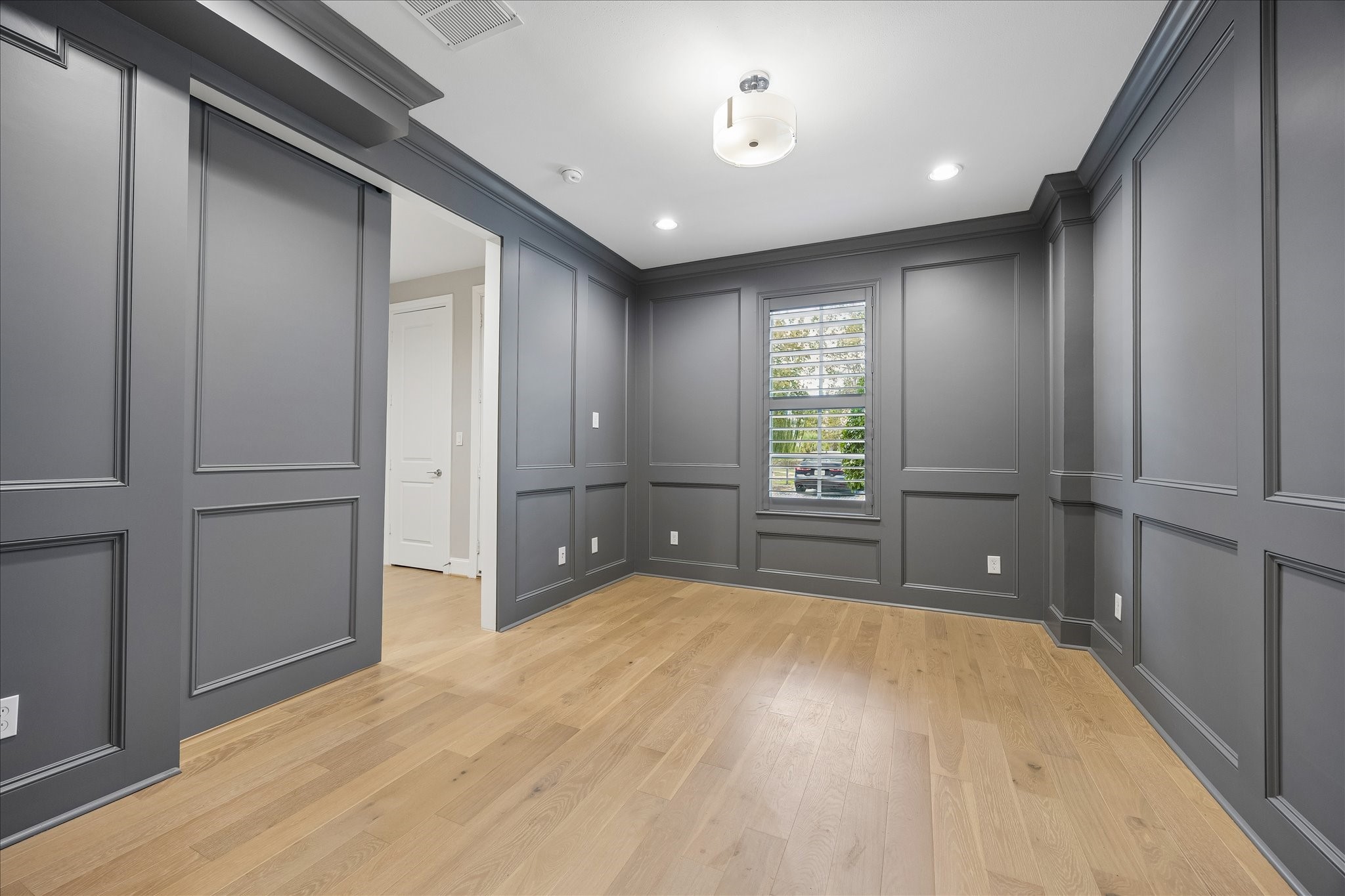 Elegant room with sophisticated dark gray wood paneled walls and warm hardwood floors, featuring recessed lighting and a modern ceiling fixture. A window with matching Plantation shutters offers natural light and a view. Perfect for an office or guest bedroom. It includes an en-suite full bath.