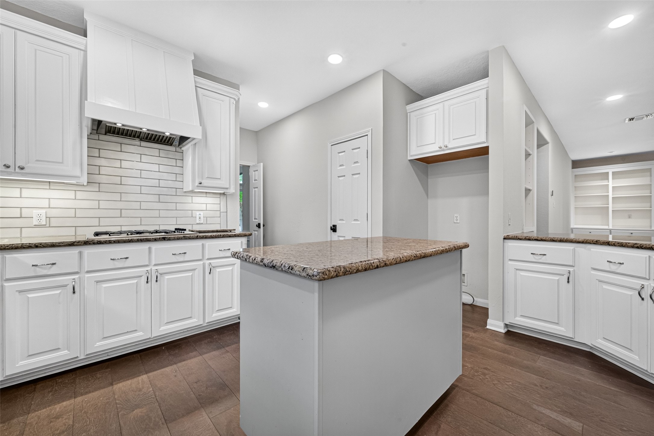 THERE'S MORE TO THIS KITCHEN THAN MEETS THE EYE! This view highlights the center island, gas cooktop & additional built-in cabinetry beyond the main cooking area. Open shelving and countertop space provide opportunities for organizing everyday essentials, displaying favorite pieces or creating a convenient beverage station. The nearby pantry adds another practical storage feature.