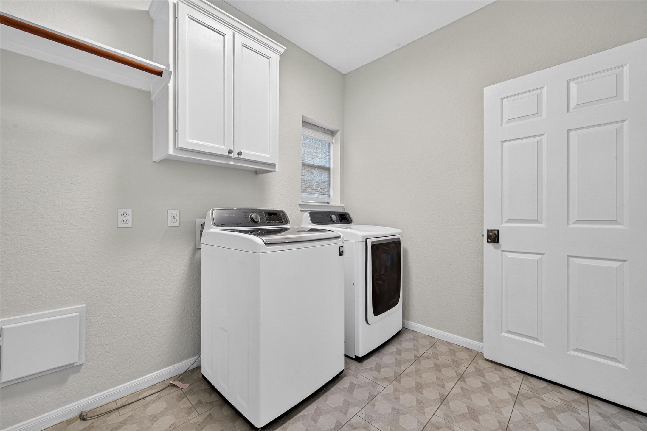 A UTILITY ROOM WITH EXTRA CONVENIENCES! This dedicated Utility Room comes complete with a full-size washer & dryer, built-in cabinetry for storing laundry supplies & a clothes rod for hanging freshly laundered garments. A window welcomes natural light, while tile flooring makes cleanup easy. There's even additional space for an upright refrigerator or freezerperfect for storing extra groceries, frozen foods or refreshments for those backyard pool parties! Practical and thoughtful design.