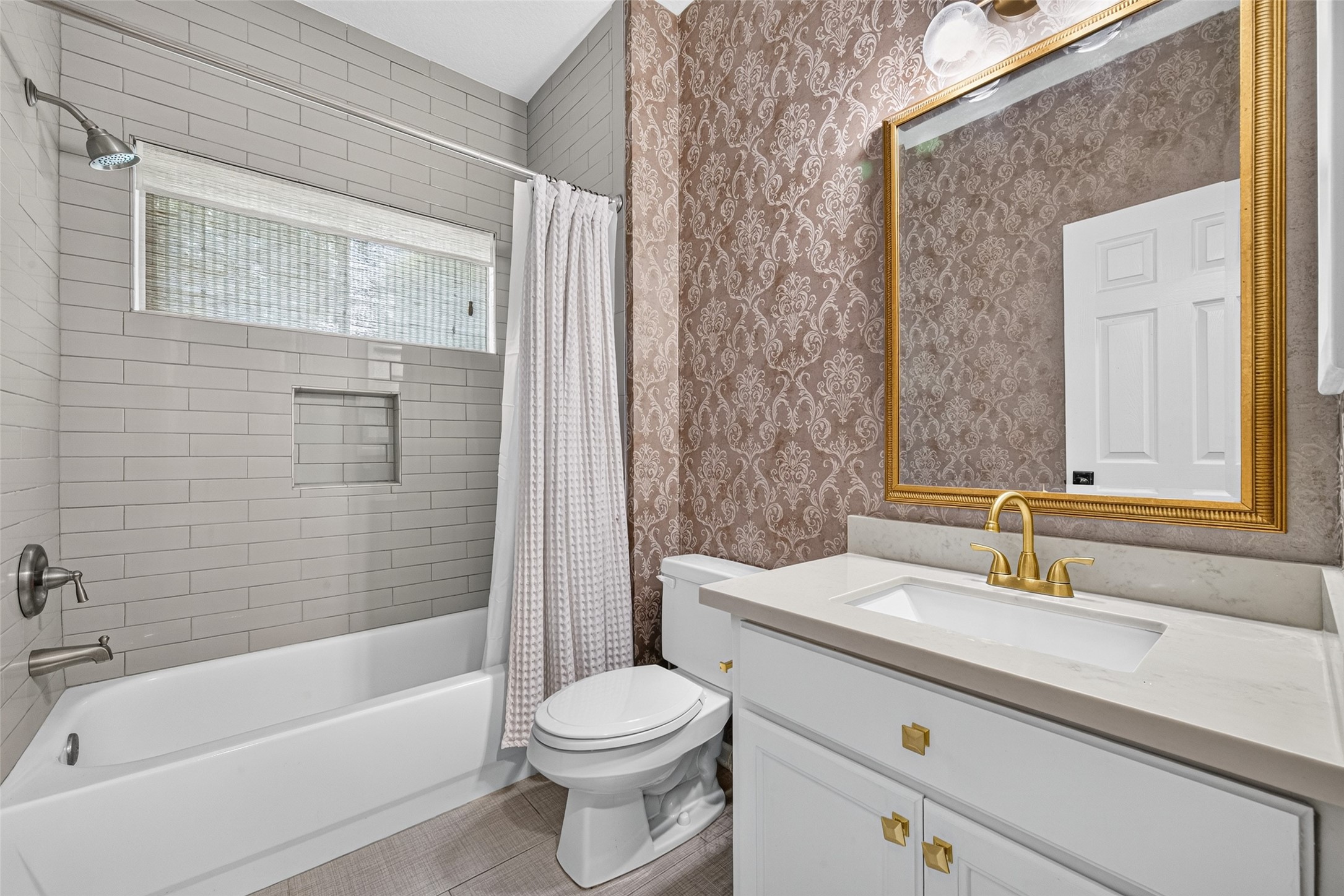 A BEAUTIFULLY APPOINTED 1ST FLOOR BATH! Conveniently located near the Study/Bedroom #4, this full bath features a tub/shower combination surrounded by white subway tile extending to the ceiling, a recessed shower niche & a window welcoming natural light. The white vanity is complemented by a light-colored countertop, brushed gold fixtures, coordinating cabinet hardware & a decorative framed mirror. Patterned wallpaper adds a touch of personality, creating a stylish space for everyday use or guests.