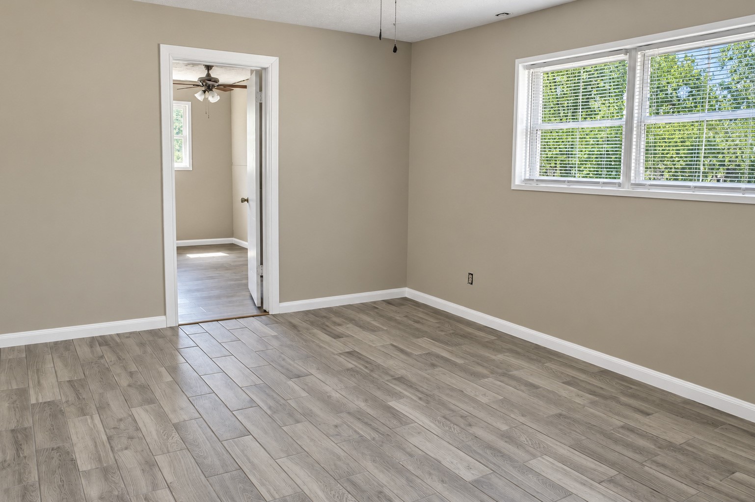 Spacious bedroom with wood-look tile flooring and abundant natural light leading into laundry room with a ceiling fan.