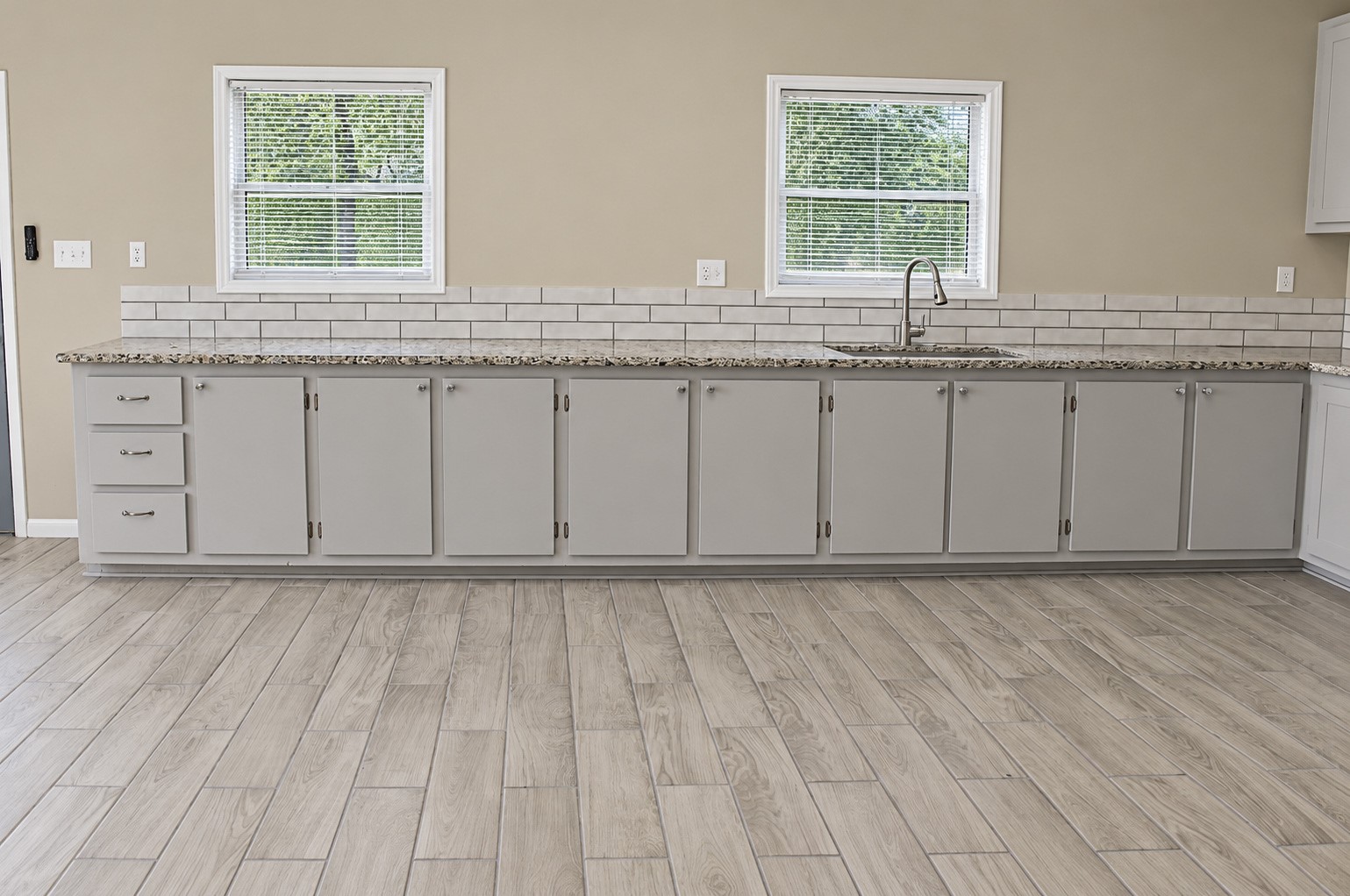 Spacious kitchen with elegant granite countertops, sleek light gray cabinetry, and a modern subway tile backsplash, complemented by warm wood-look tile flooring.
