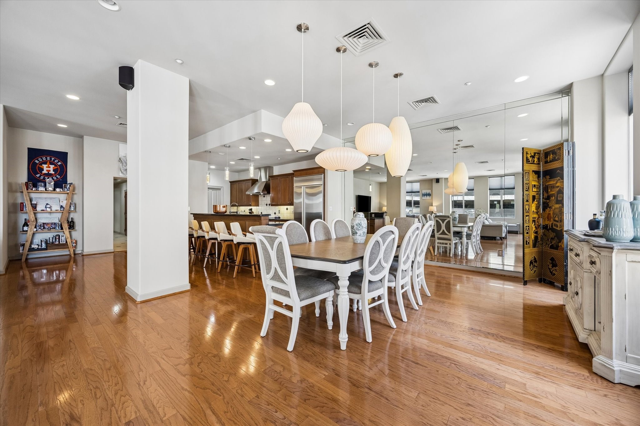 Spacious open-concept dining area with elegant hardwood floors, large mirrored wall, and modern pendant lighting, seamlessly flowing into a contemporary kitchen with stainless steel appliances and a breakfast bar.