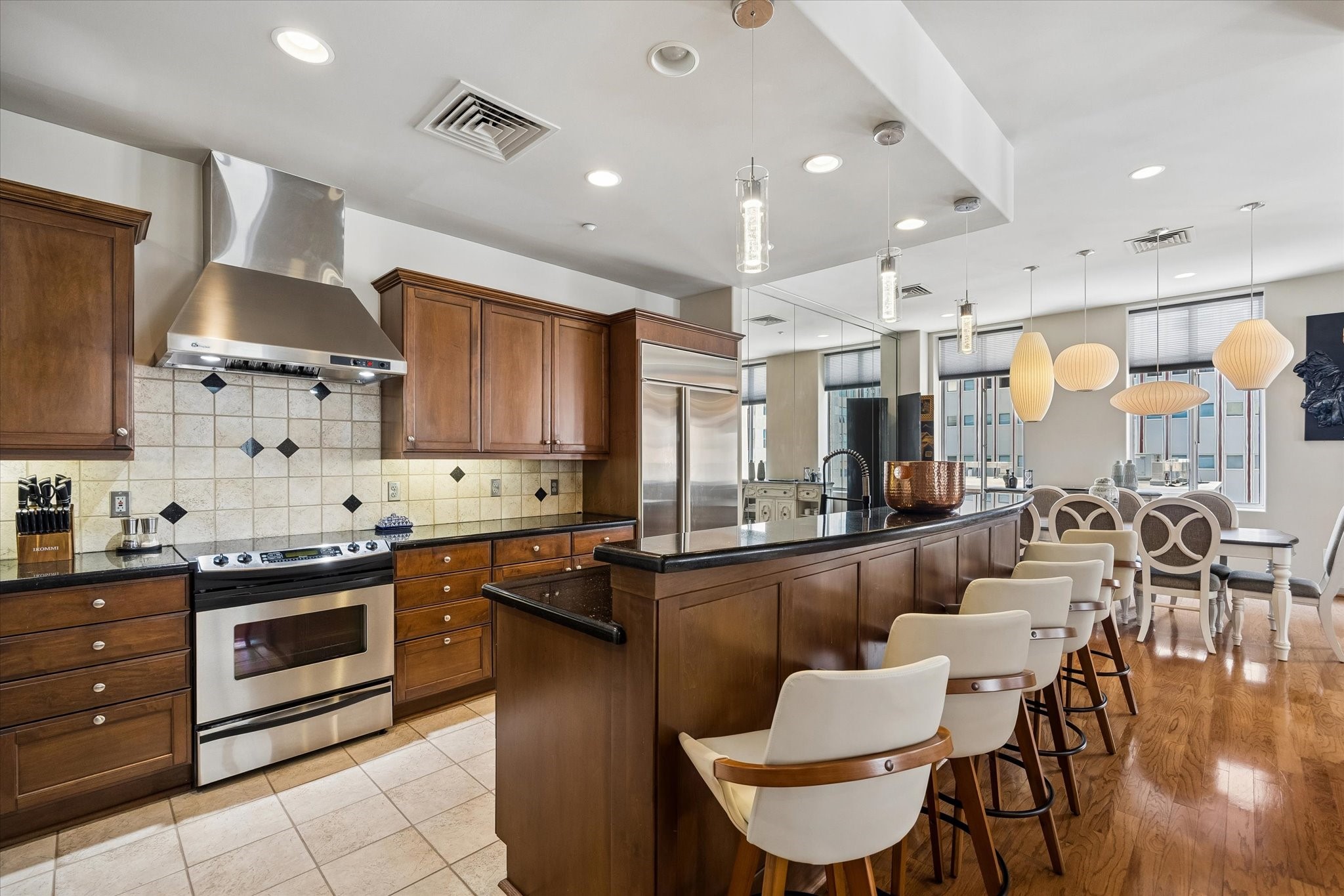 Elegant kitchen with rich wood cabinetry, stainless steel appliances, and a stylish tile backsplash. The open layout includes a breakfast bar with chic seating, flowing seamlessly into a bright dining area enhanced by modern pendant lighting.