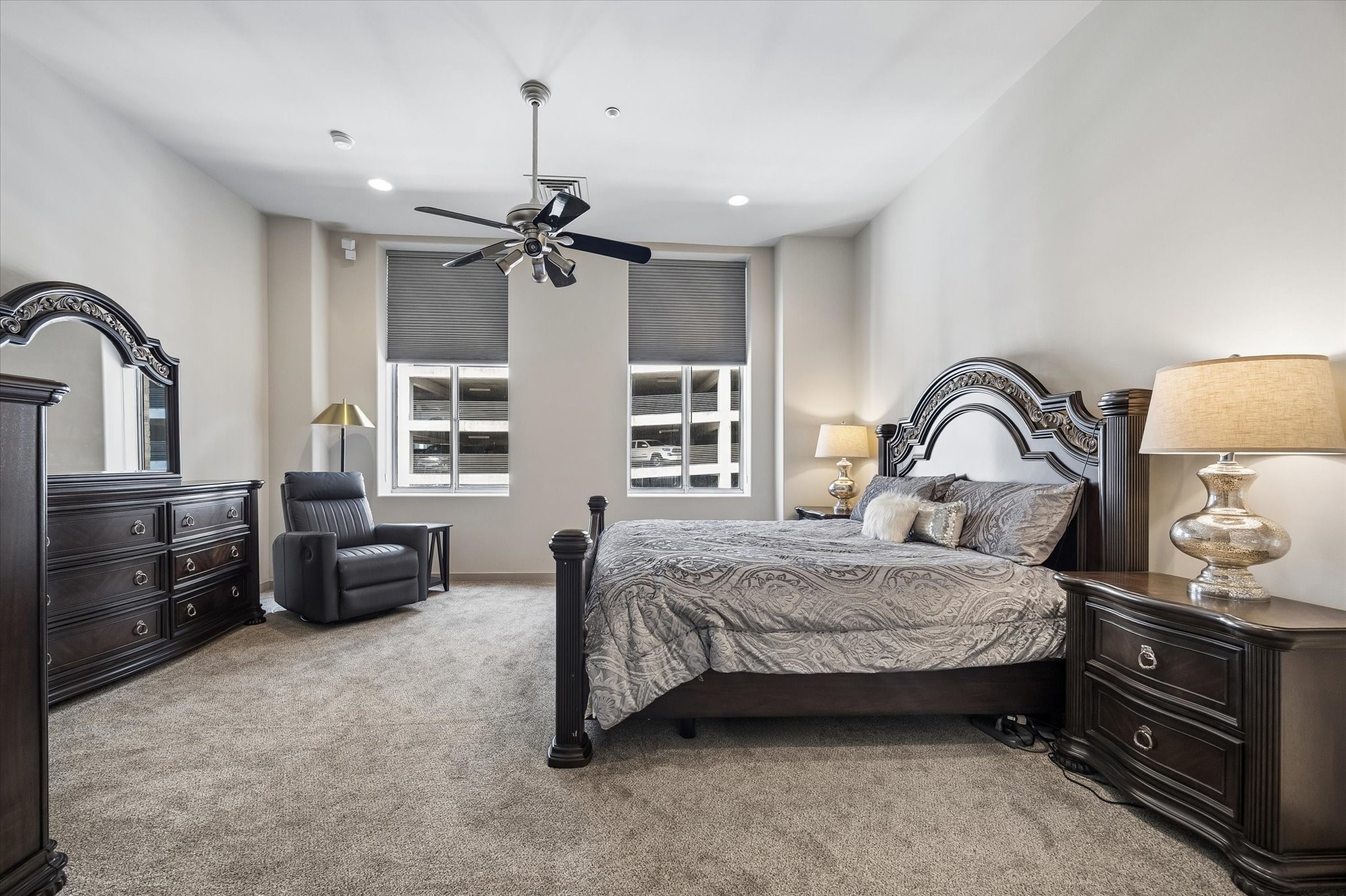 Spacious and elegant primary bedroom with plush carpeting, featuring a luxurious bed set, matching dark wood furniture, and modern ceiling fan, all illuminated by ample natural light from large windows.