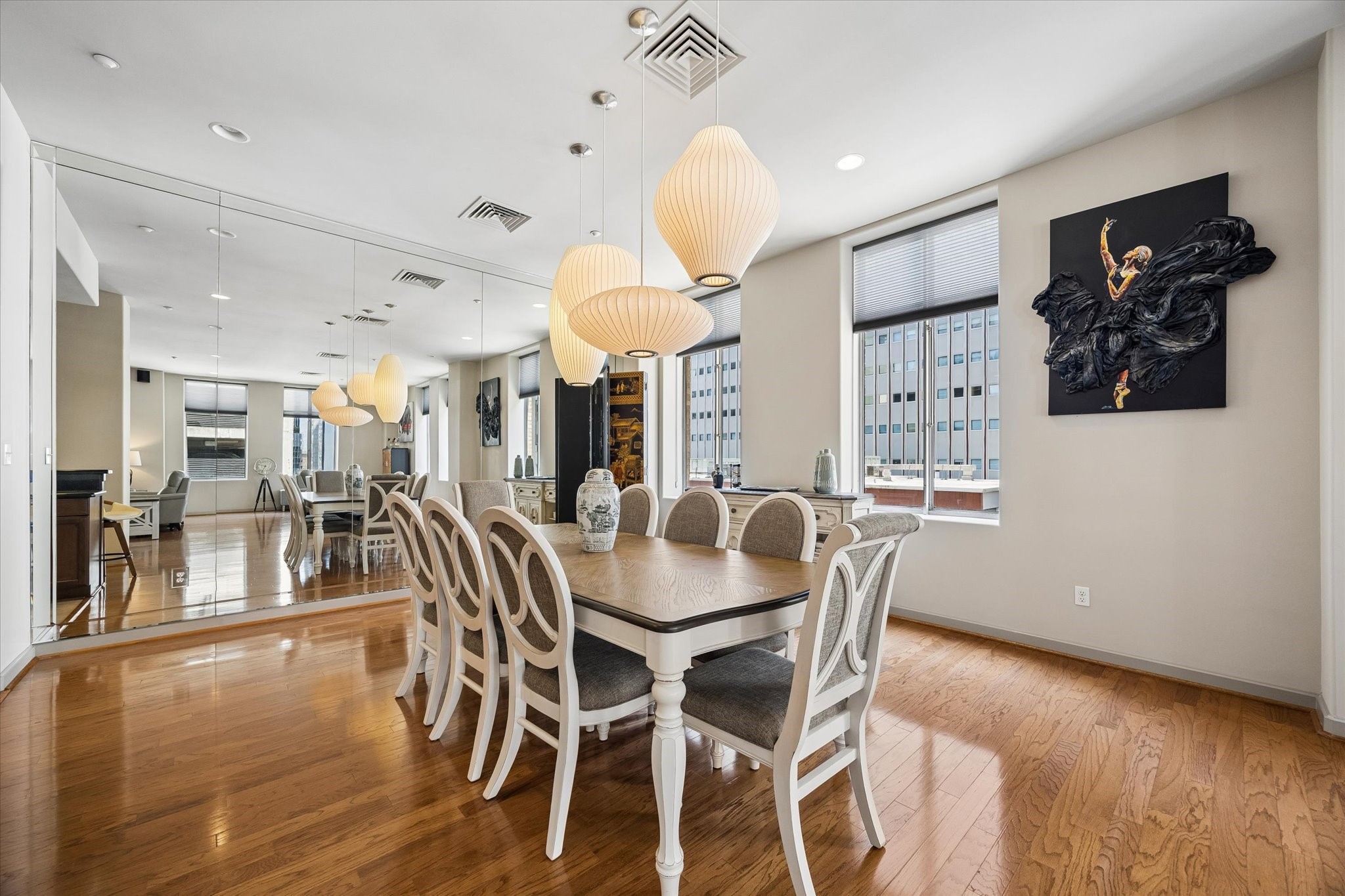 Elegant dining area featuring a spacious table with upholstered chairs, accented by stylish pendant lighting and a large mirrored wall that enhances the room's brightness and sense of space.